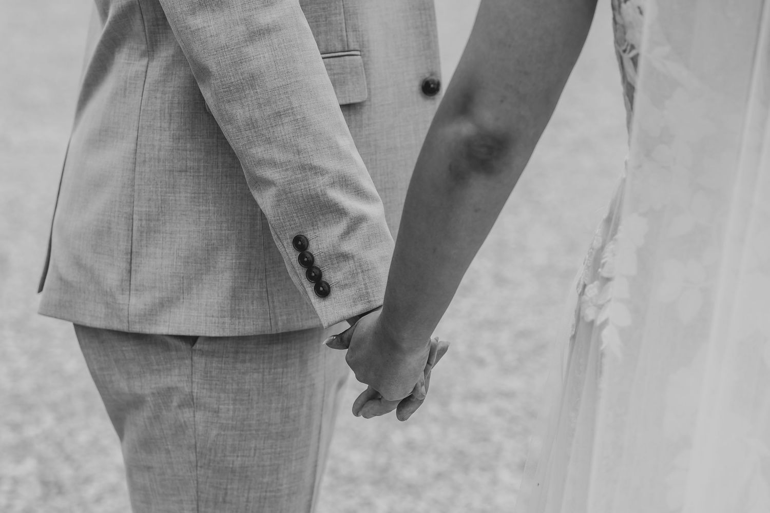 Black and white close-up of newlyweds holding hands, showing wedding attire details.