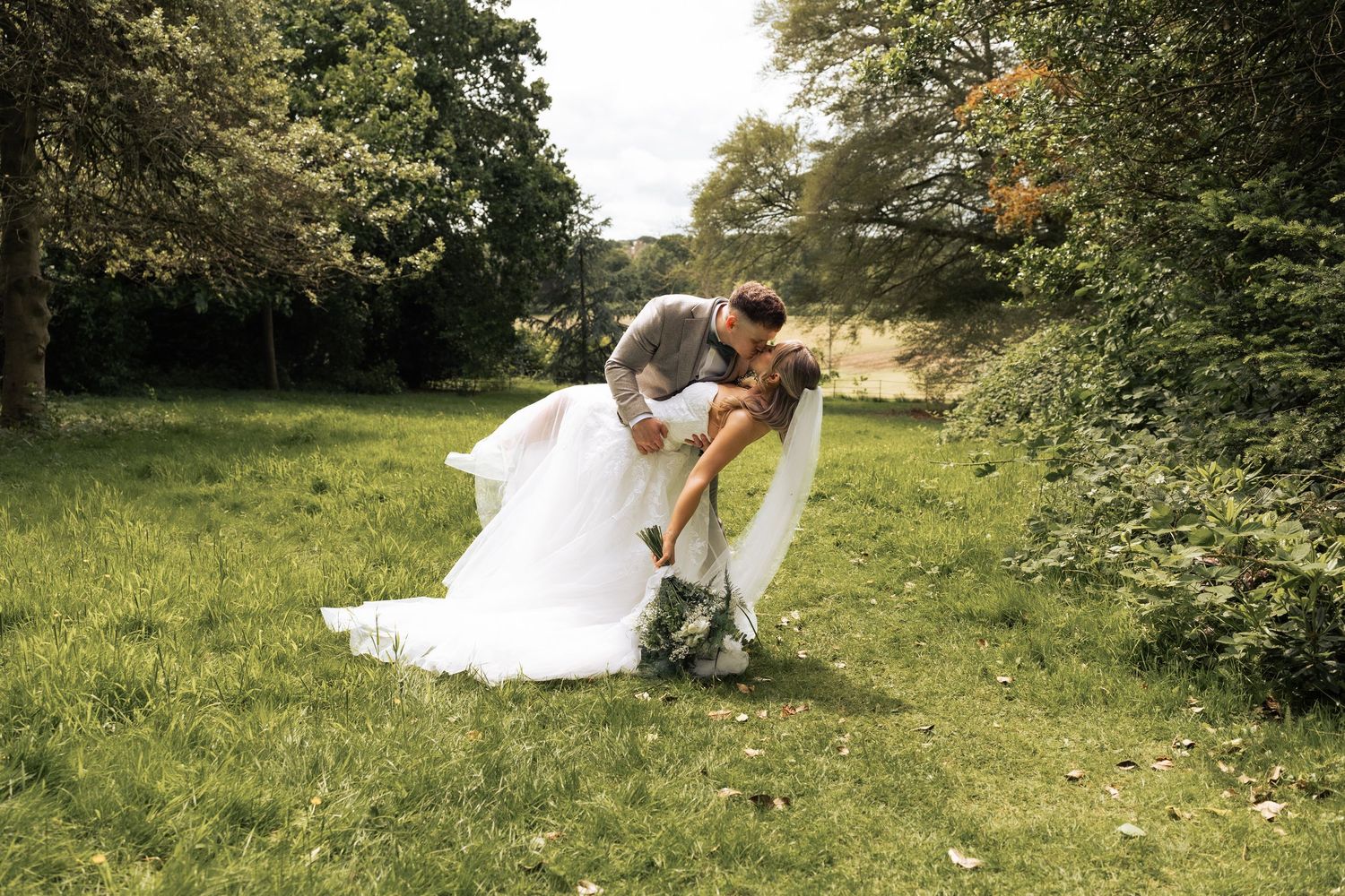Groom dipping bride for a romantic kiss in a lush green garden on their wedding day.