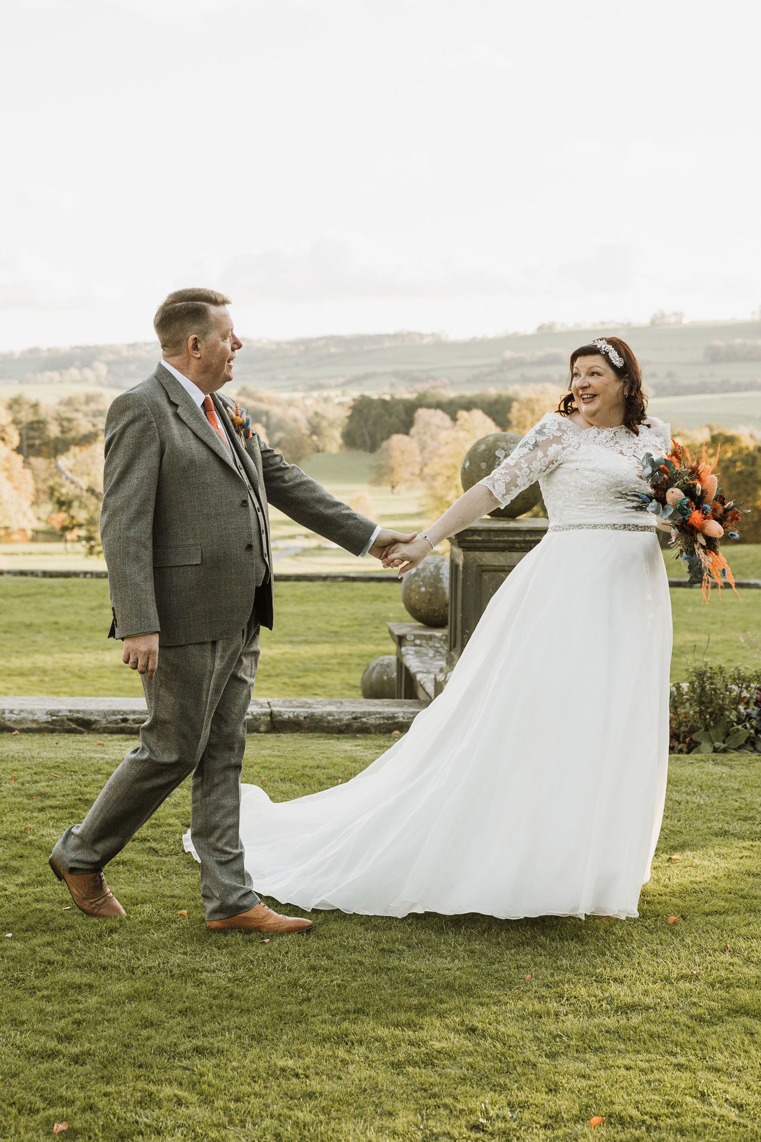 A bride in white lace gown holds hands with groom in gray suit, dancing outdoors with scenic countryside backdrop.