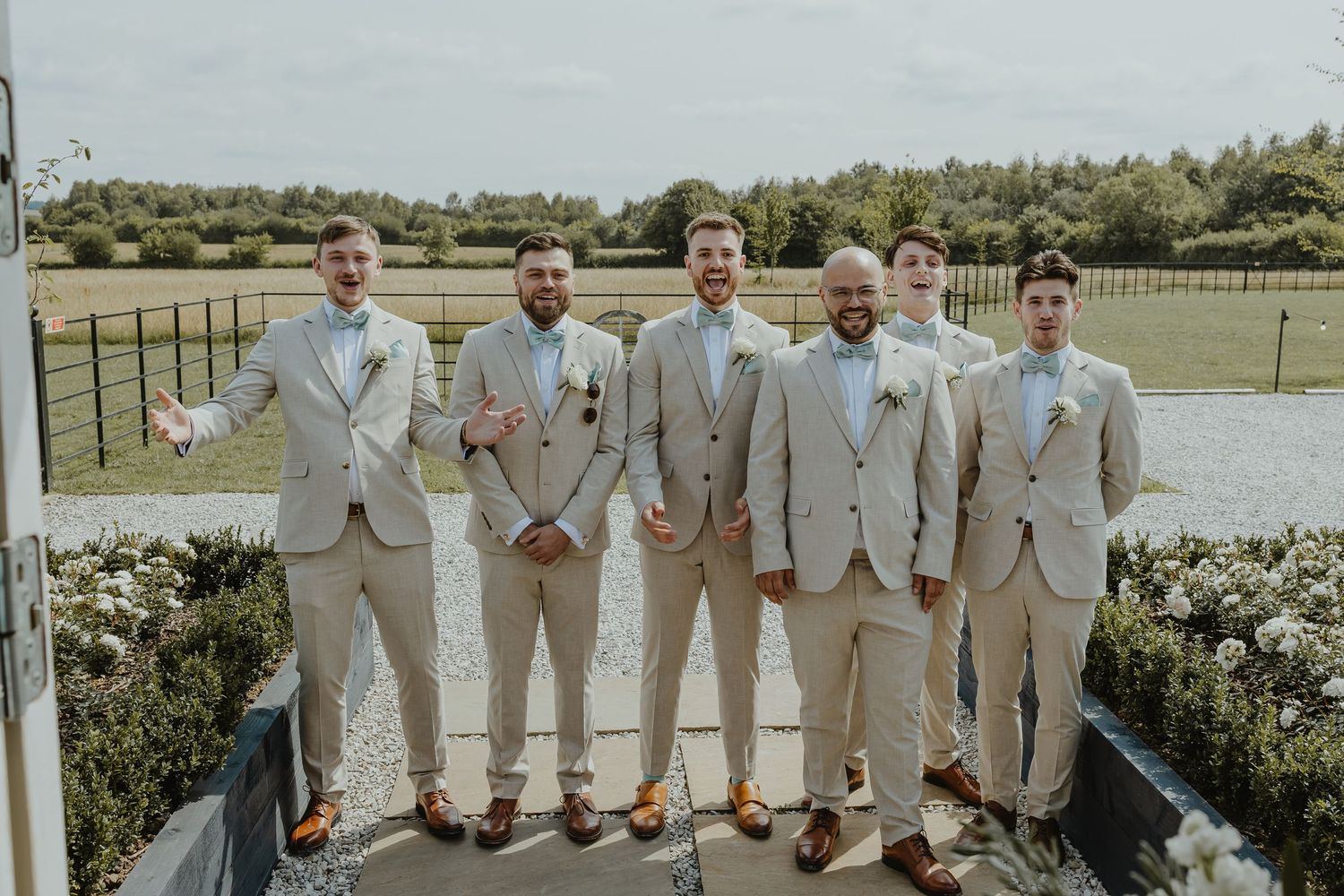 Wedding party members in tan suits and light blue dresses pose together outside.