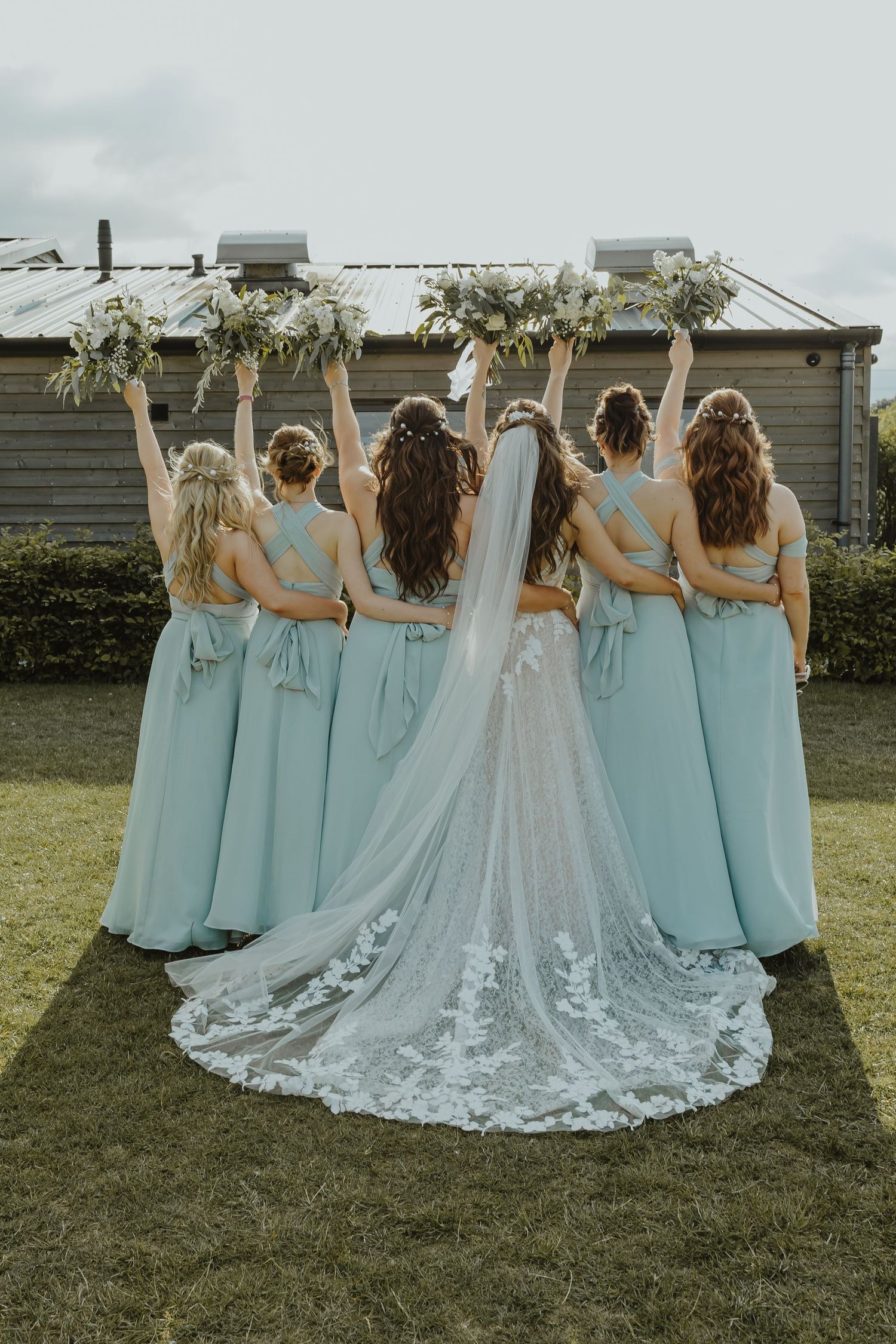 Bridesmaids in blue dresses pose with bride showing off lace train of wedding dress from behind.