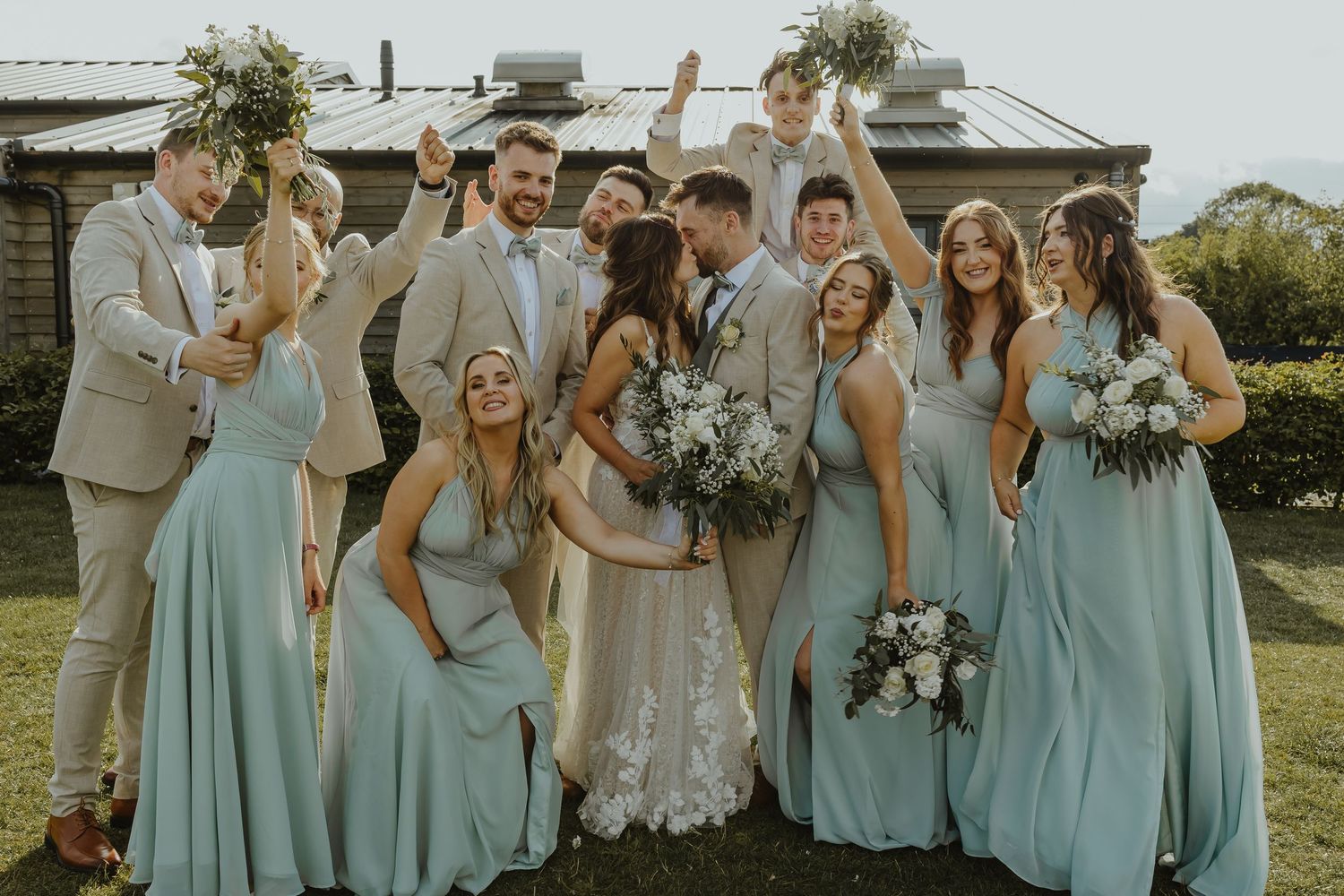 Wedding party in mint green bridesmaid dresses and beige suits celebrate joyfully outdoors.