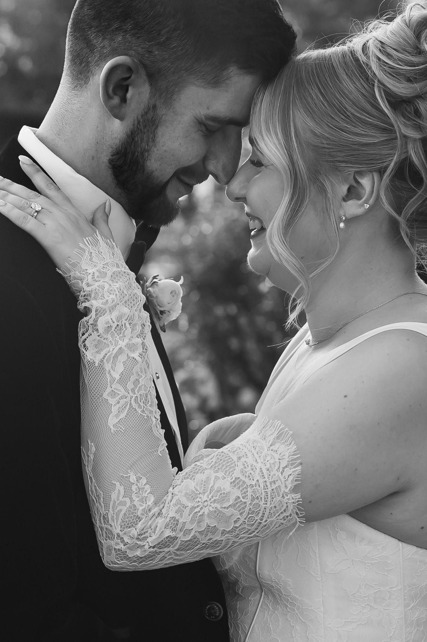 Black and white wedding photo of smiling bride in lace gown and groom touching foreheads tenderly outdoors.