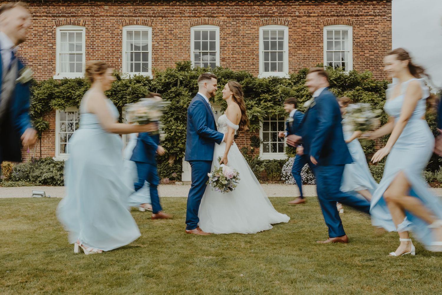 Wedding party running and playing on grass in front of historic brick building.