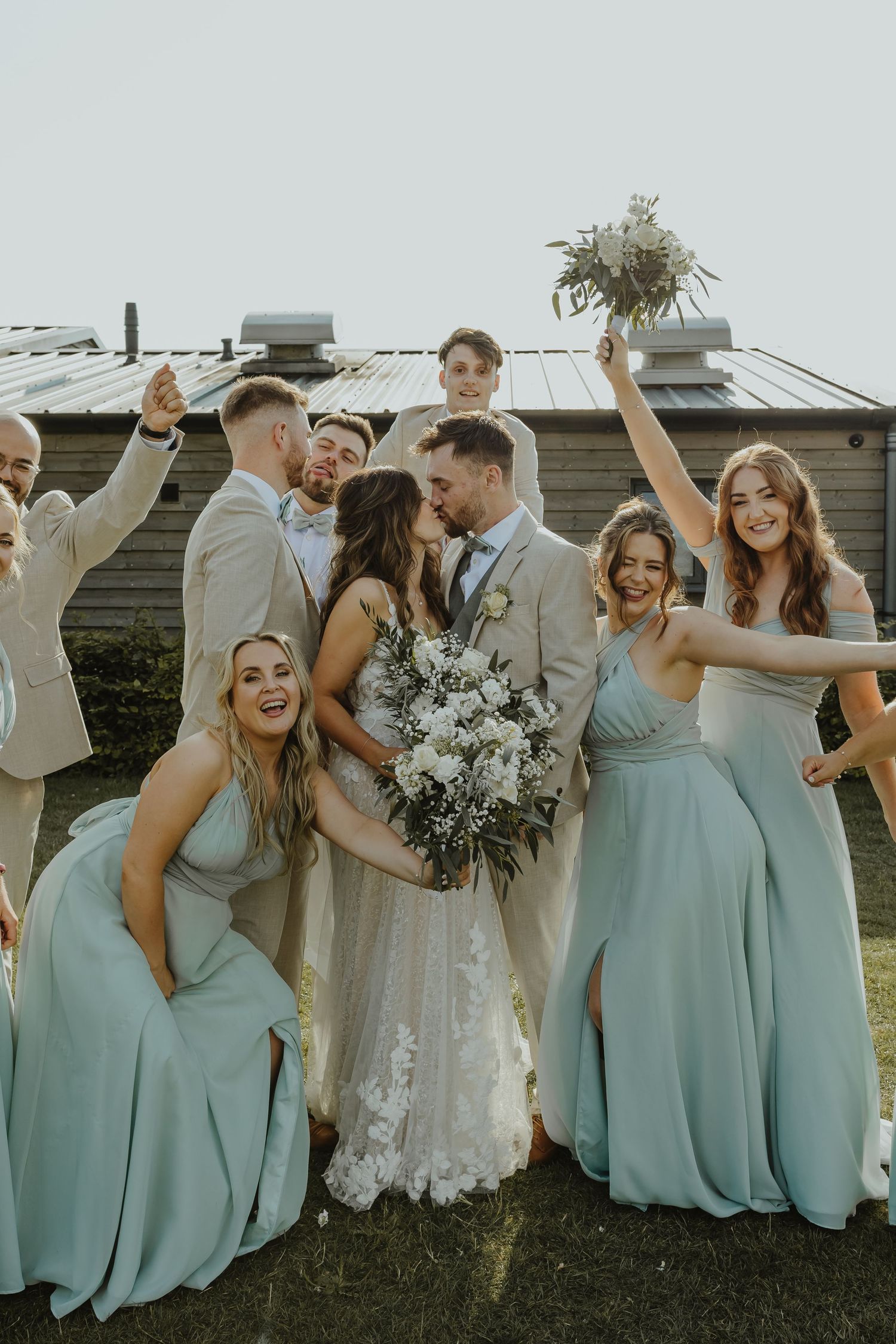 Wedding party celebrates joyfully outside rustic venue with bridesmaids in blue dresses.