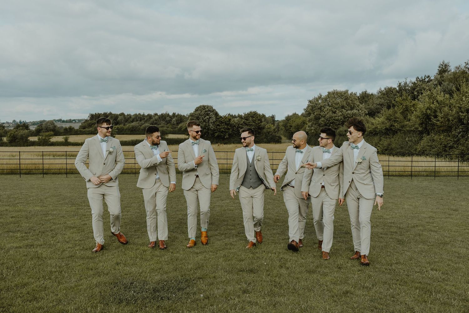 Group of groomsmen in light suits walking together across grass field under cloudy sky.