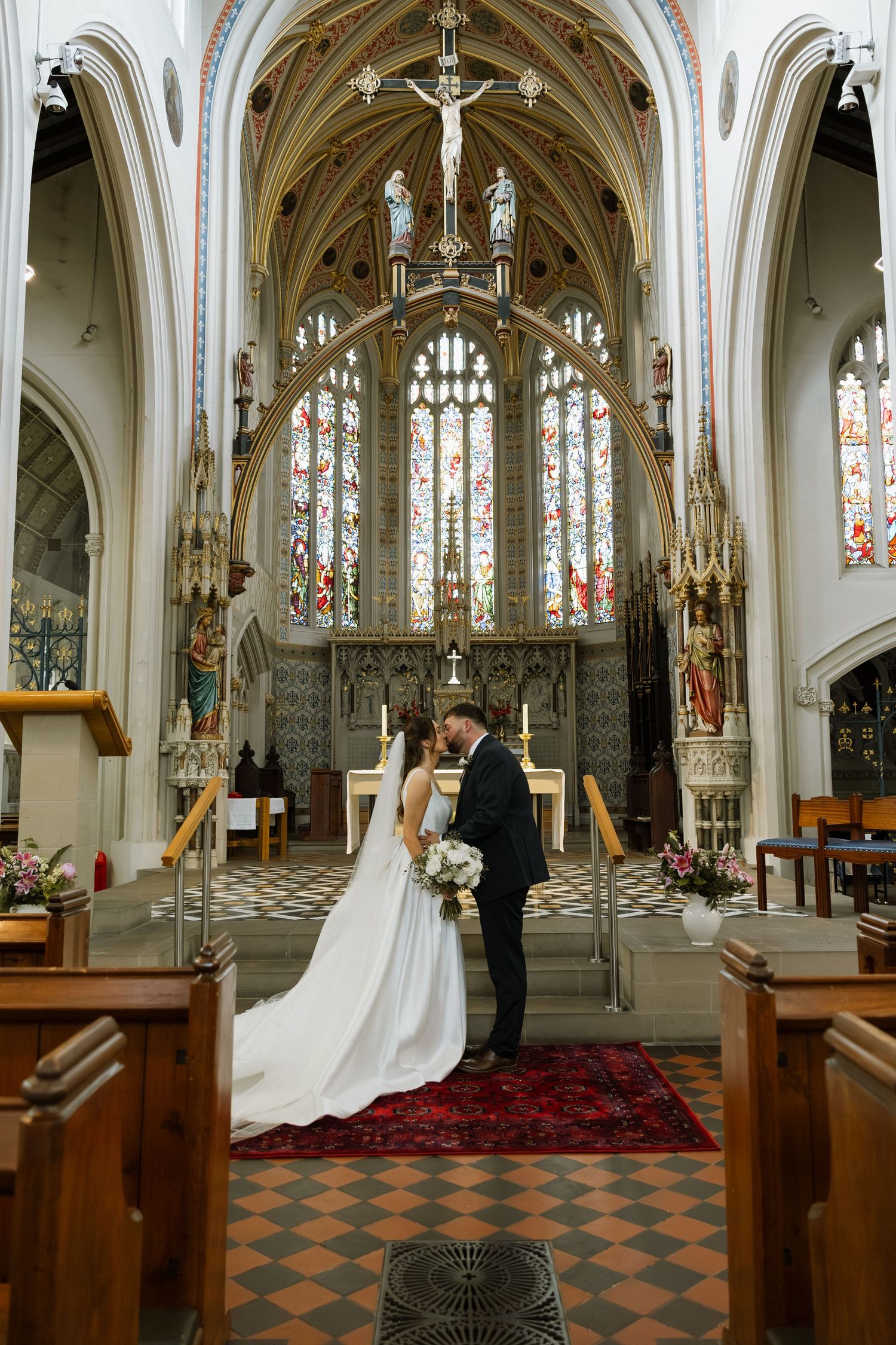 Bride and groom share a kiss at the altar of an ornate Gothic church with stained glass windows.