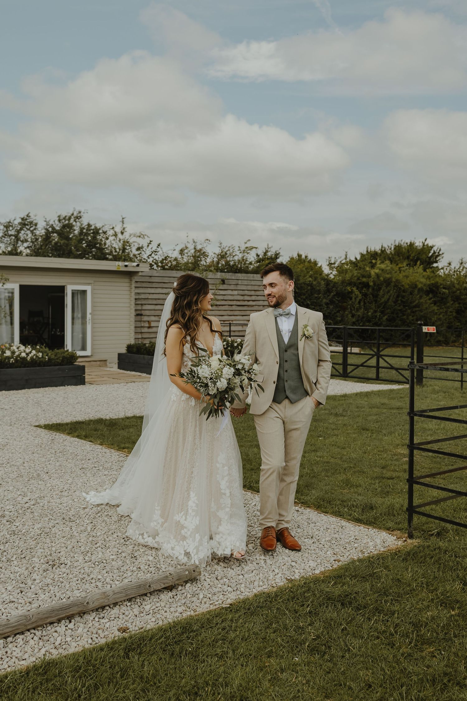 Newlyweds pose together on a gravel path outside a rustic venue.