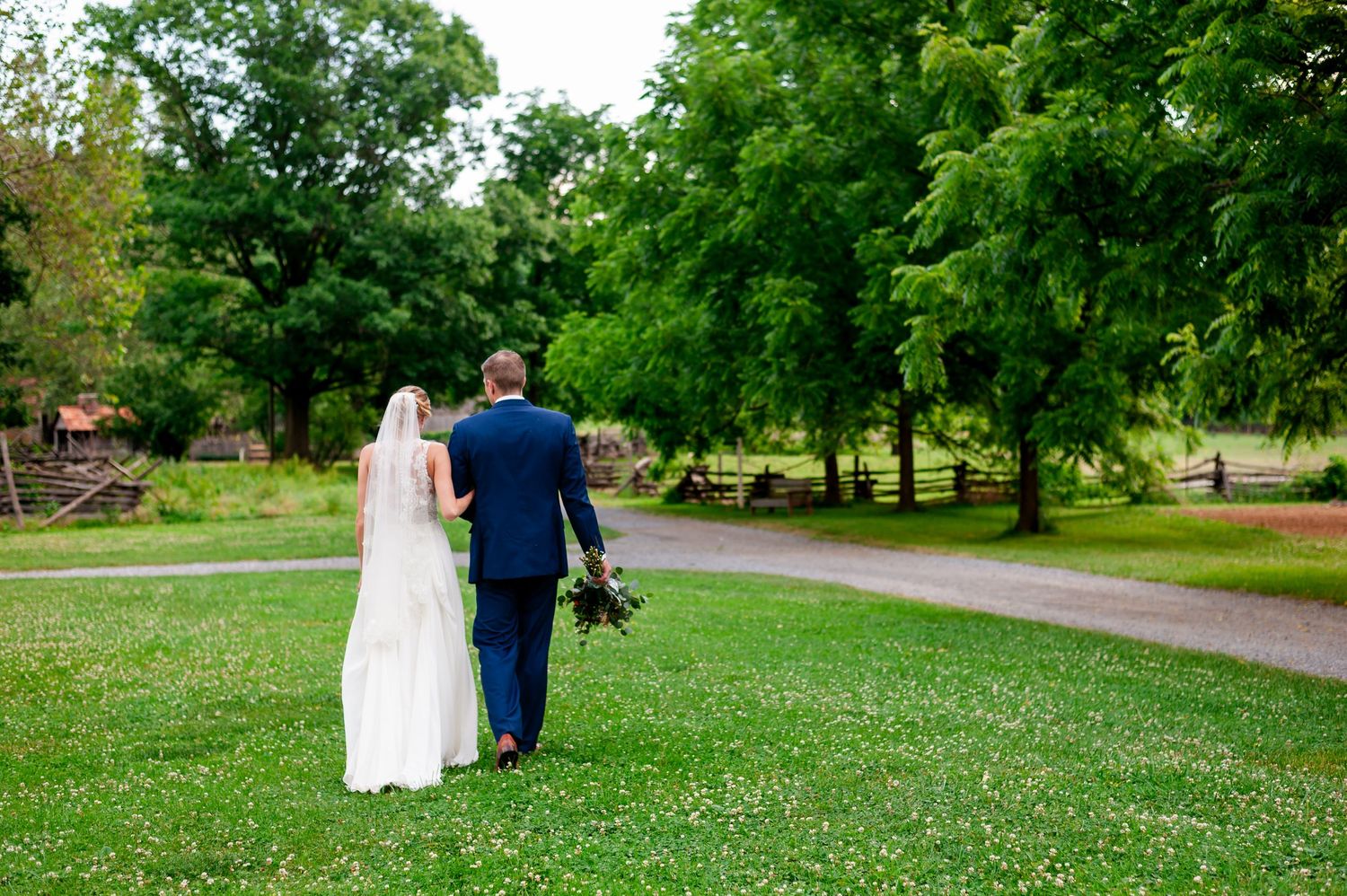 Bride and groom walk together on a park path surrounded by lush summer greenery.