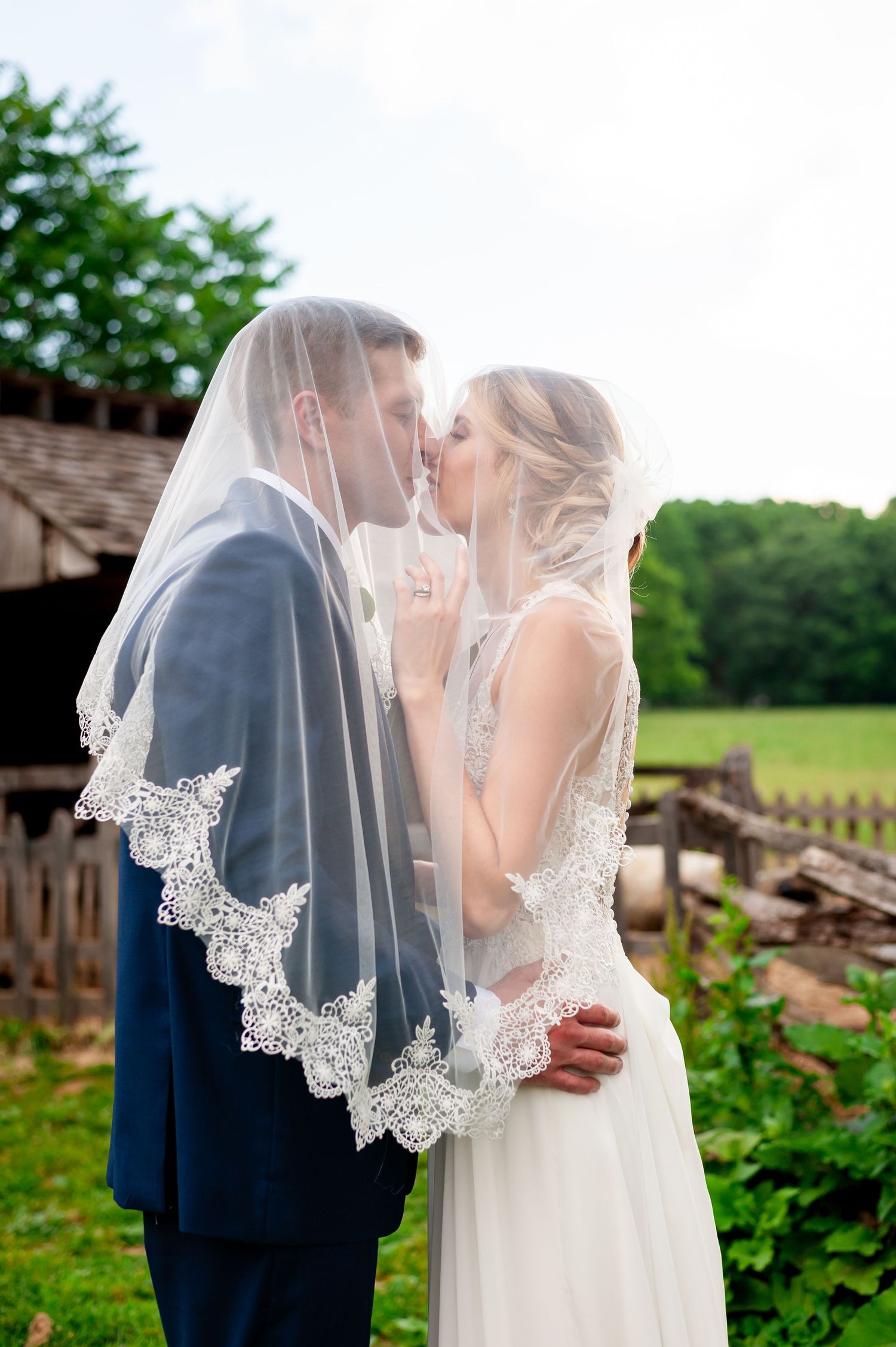 Bride in lace veil kisses groom in navy suit amid garden setting.