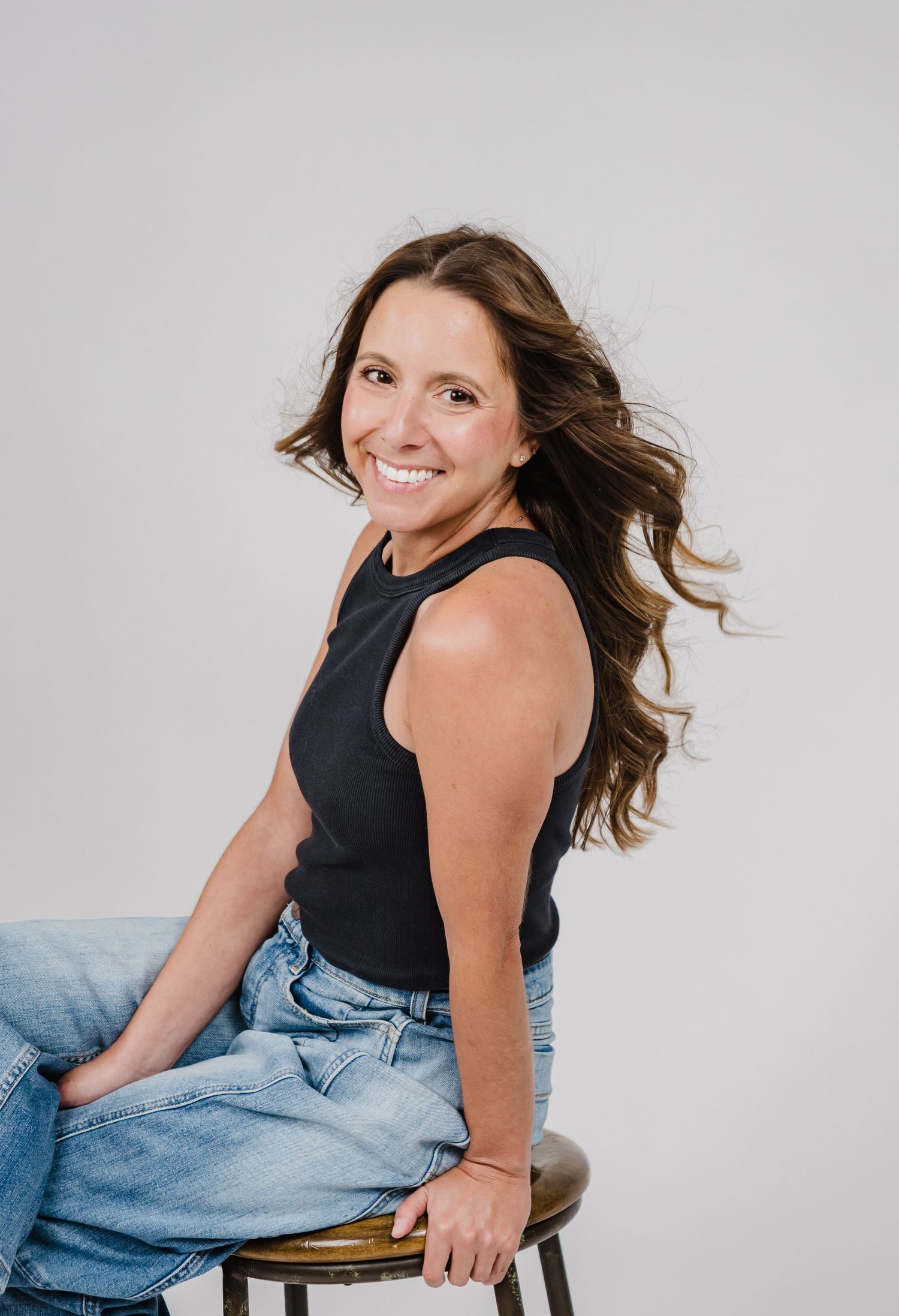 Portrait of a person in a black sleeveless top and light denim jeans sitting casually on a stool against a gray background.