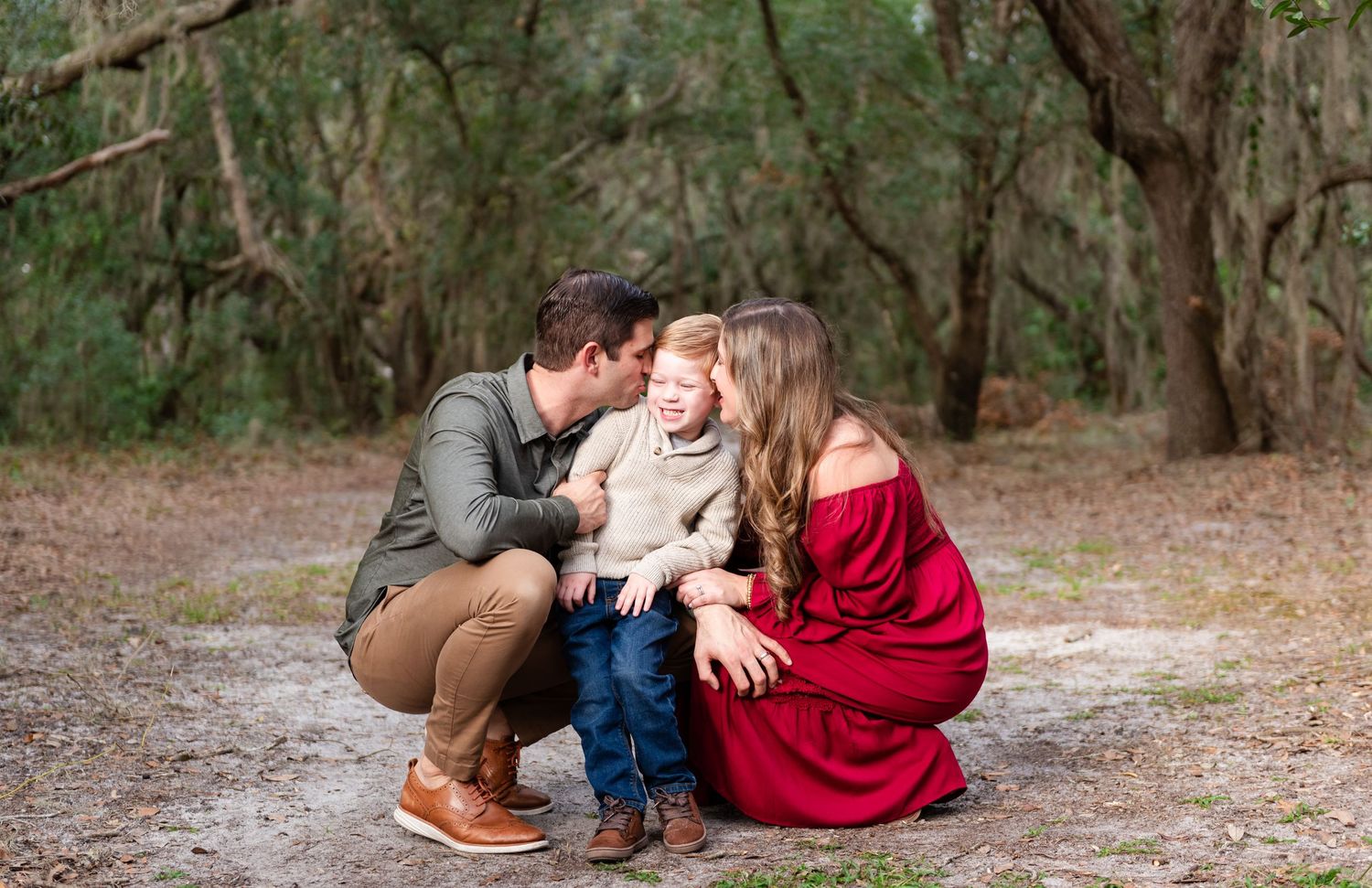 A couple in casual attire shares a tender moment with their toddler on a wooded sandy path.