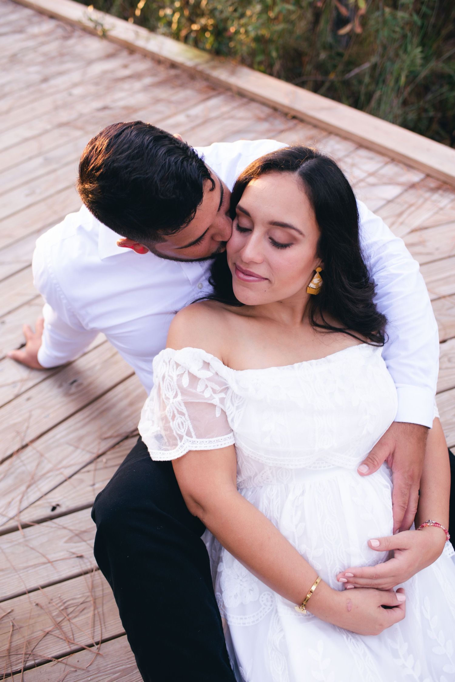 Romantic maternity photo of couple embracing on wooden deck while sharing intimate moment in white lace dress.