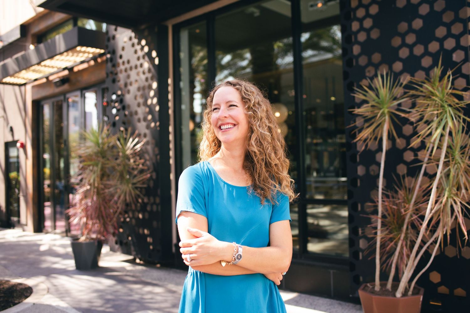 Someone with curly hair wearing blue laughs joyfully outside a commercial building.