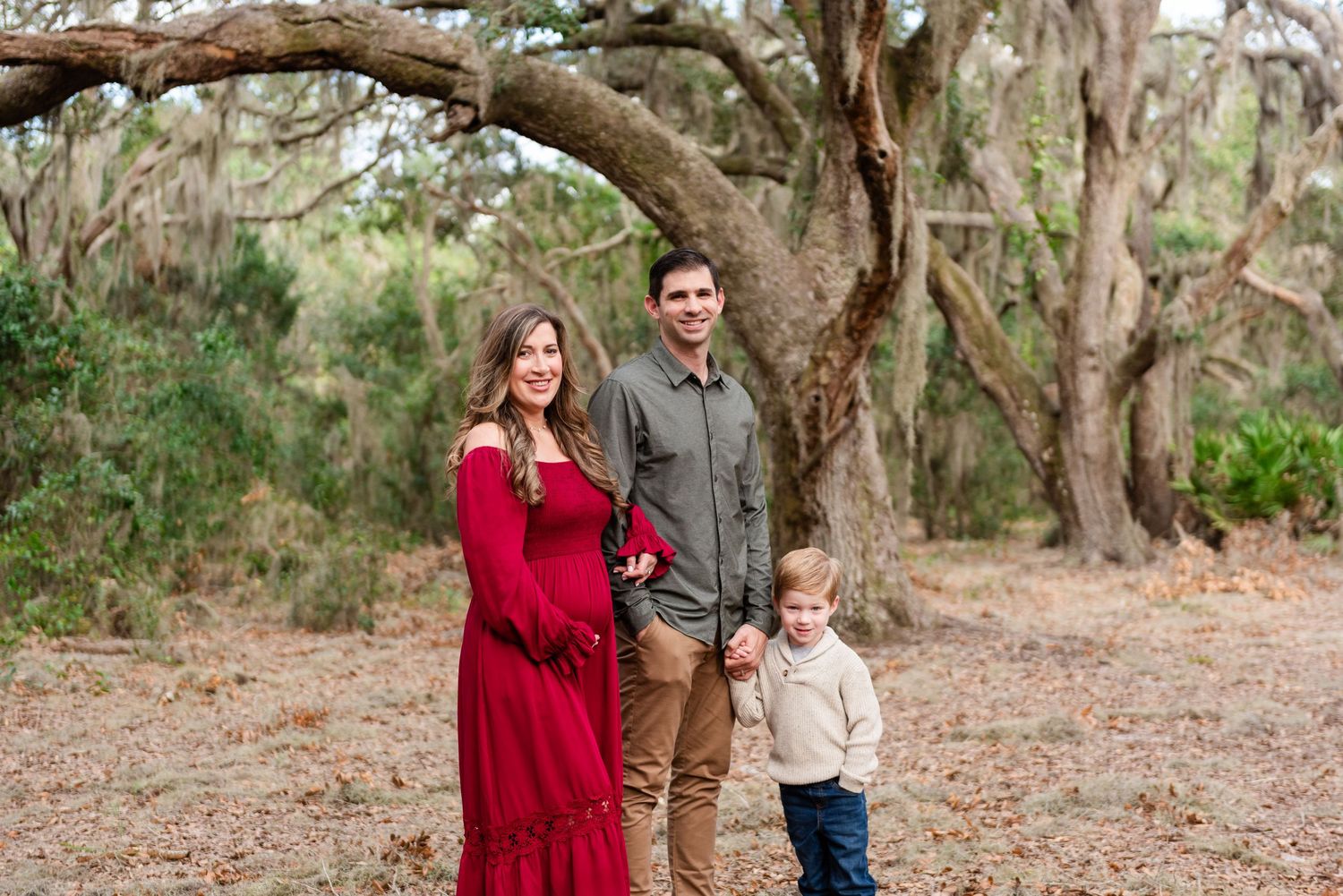 Three people stand together on a dirt path surrounded by large oak trees with hanging moss in a natural outdoor setting.