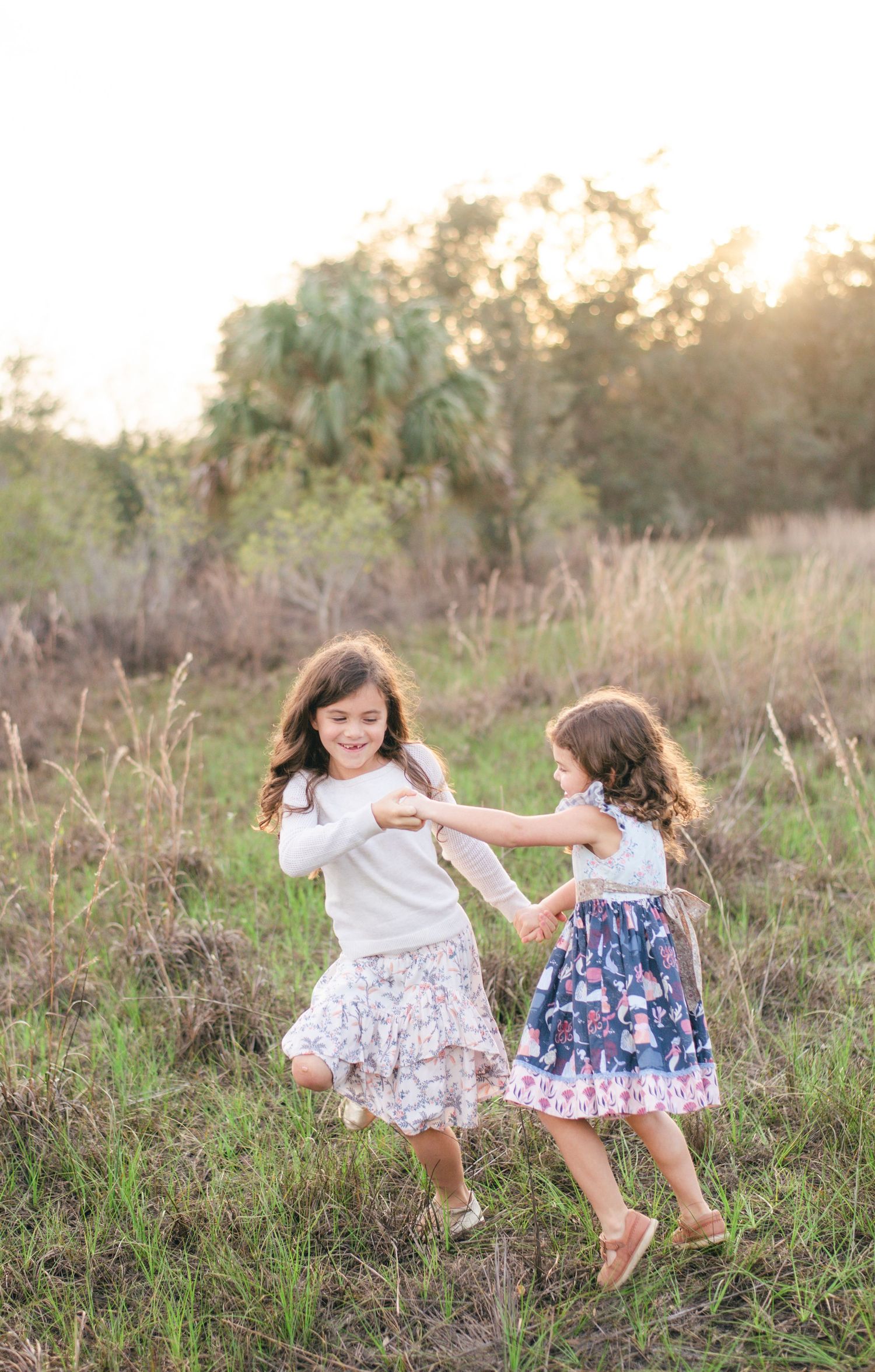 Two young children in white and floral dresses dance and play together in a grassy field at sunset.