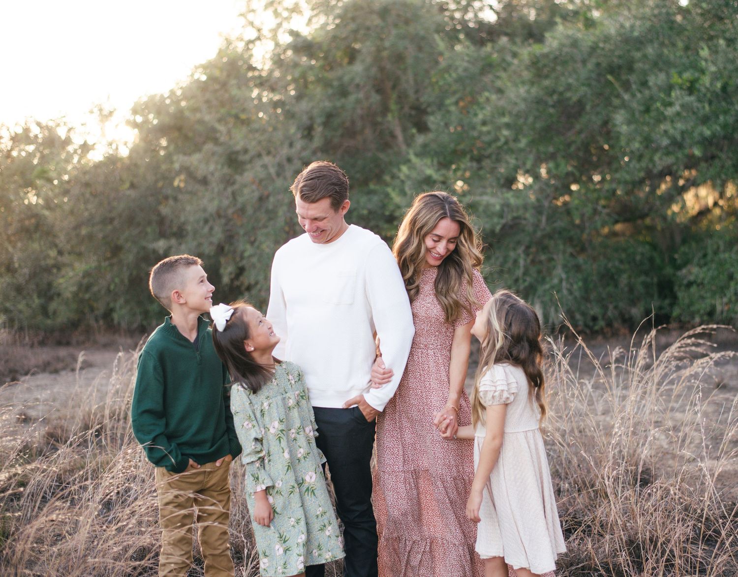 A family stands together in a golden field at sunset wearing coordinated white, green and pink outfits.