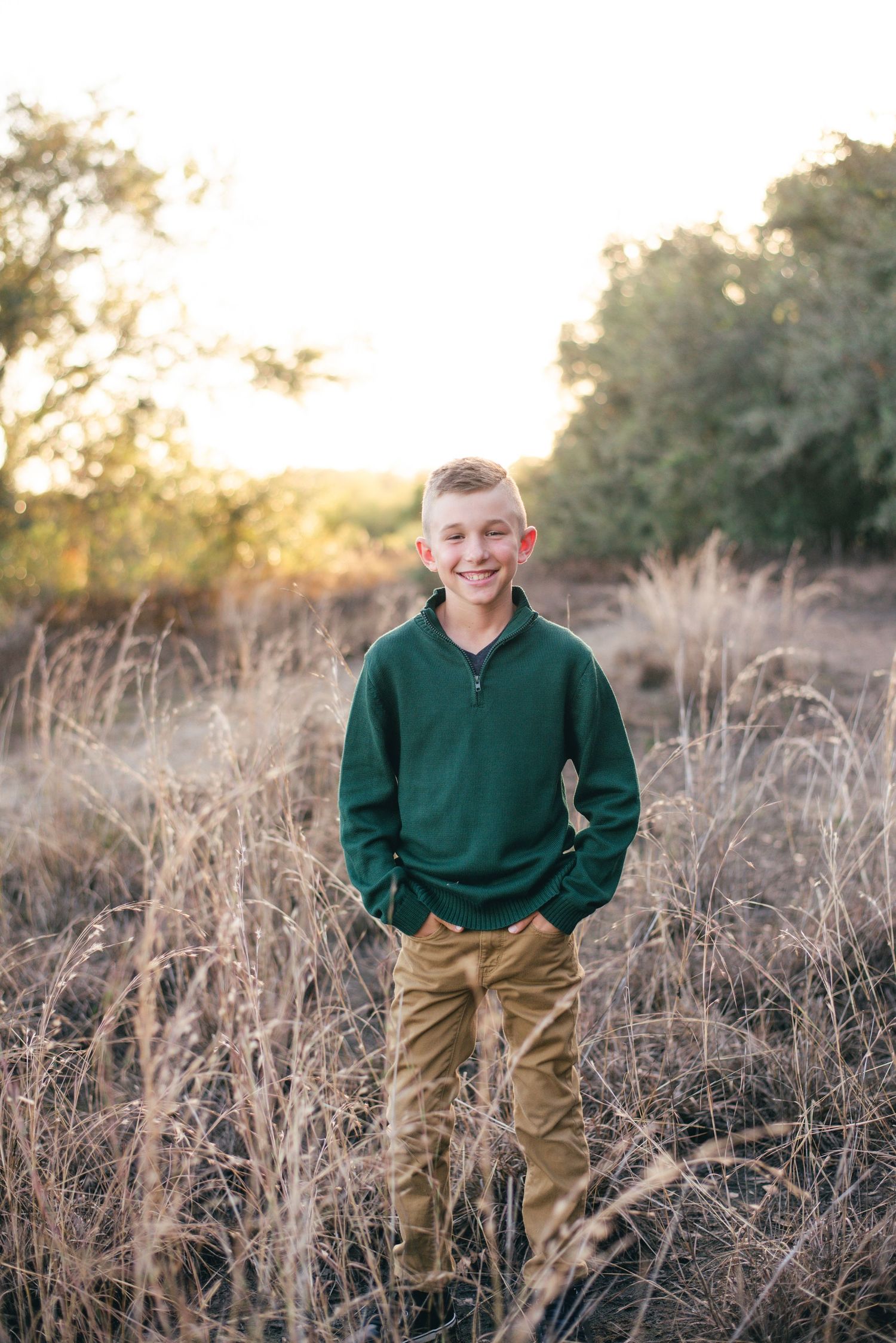 Young person stands in tall dried grasses wearing a forest green sweater and khaki pants during golden hour.