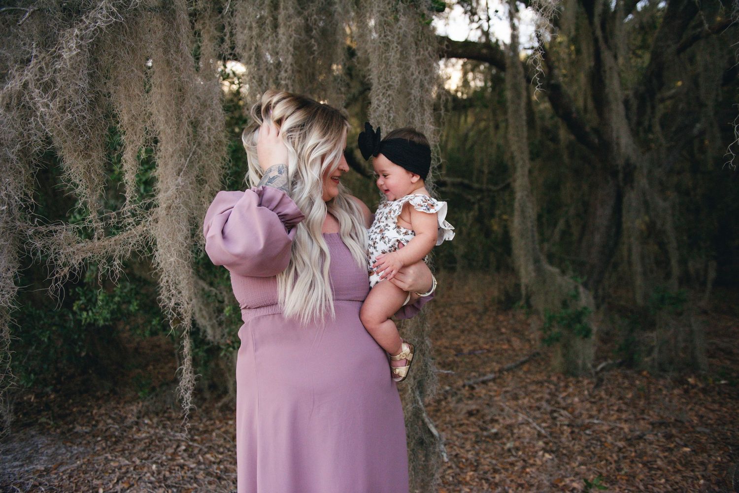 Figure in mauve dress holding infant outdoors among Spanish moss-draped trees.