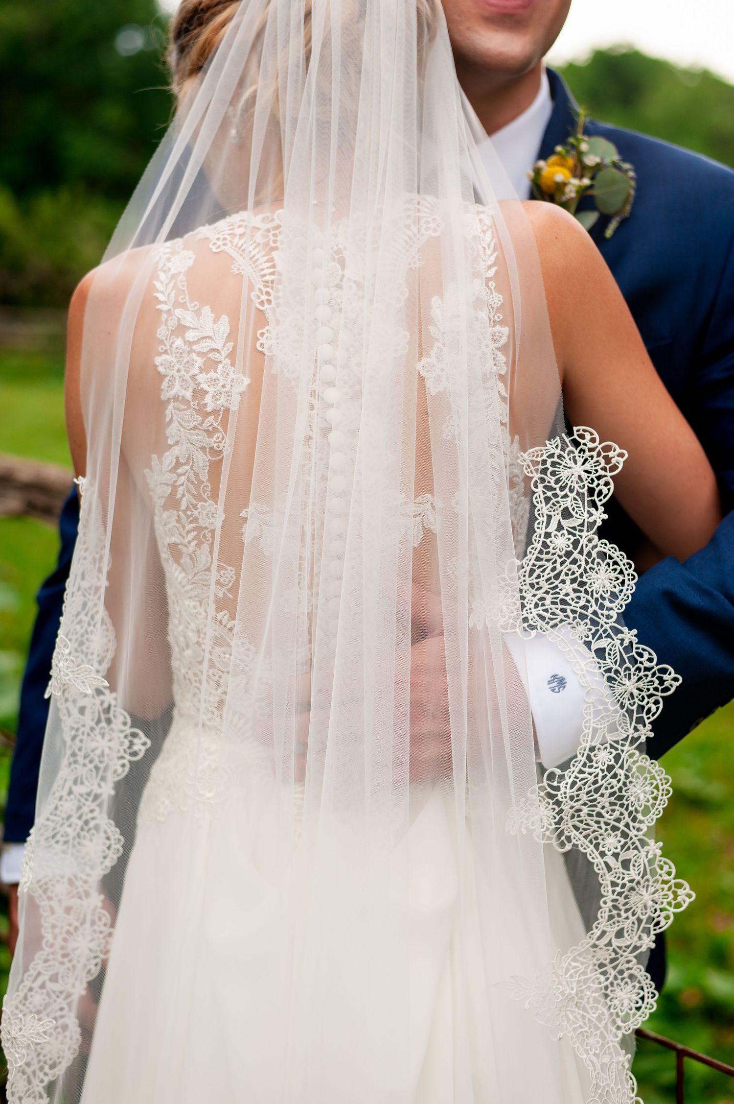 Close-up detail of an ornate lace wedding veil and white dress against a navy suit.