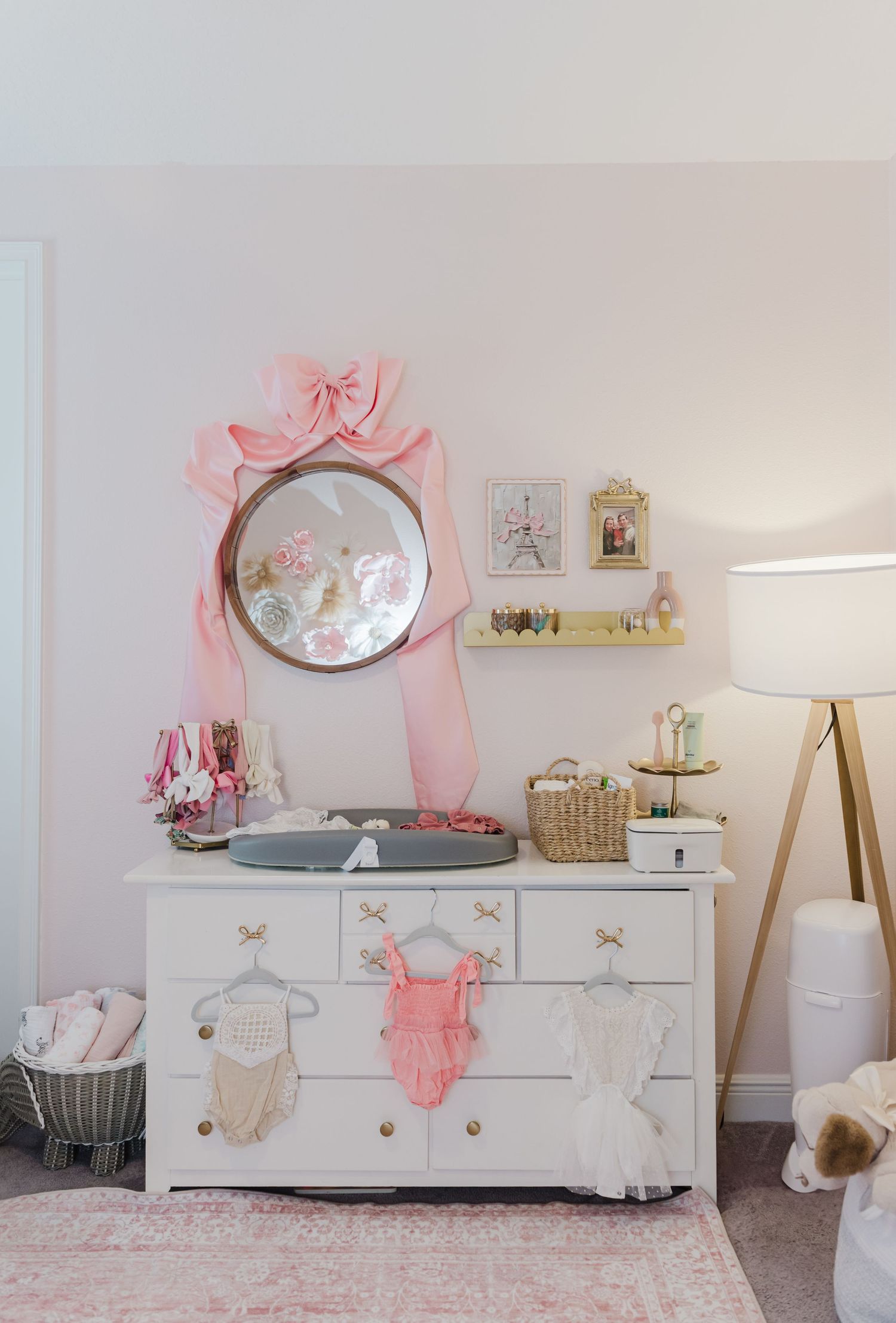 White dresser with mirror in a pink nursery decorated with baby clothes and accessories.