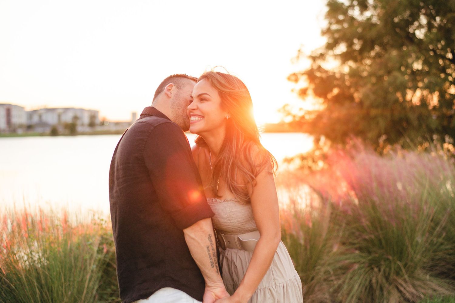 A couple shares an intimate sunset moment by the water surrounded by tall grasses and warm golden light.