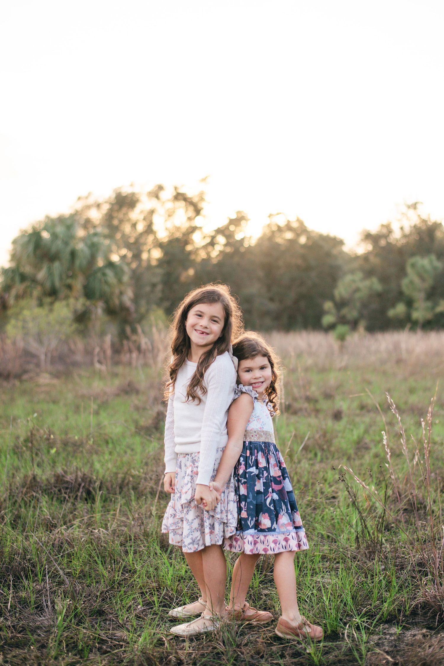 Two people stand together in a sunlit meadow with tall grass during golden hour.