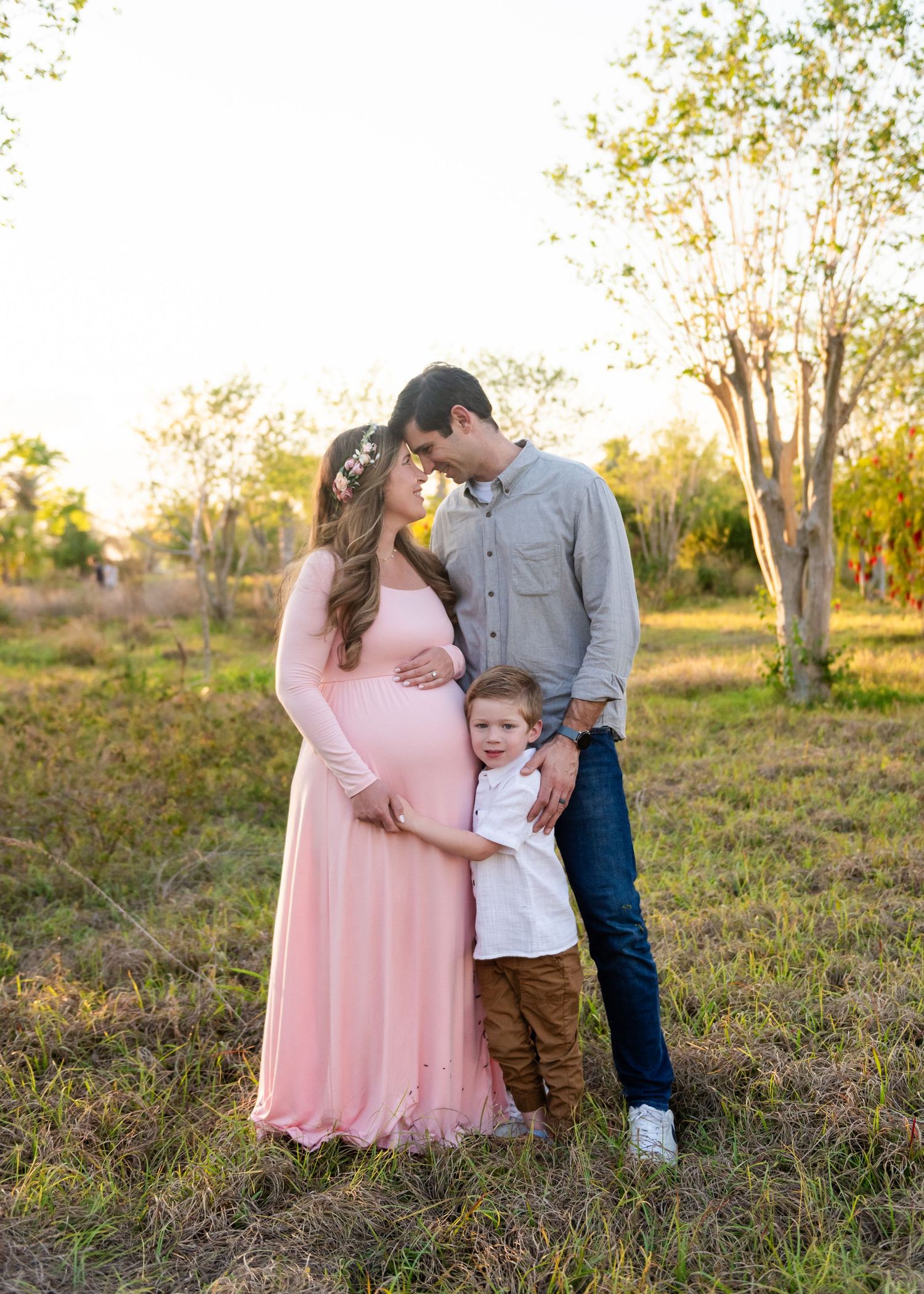 A pregnant woman in a pink dress stands with a young child in a field at sunset while wearing a flower crown.