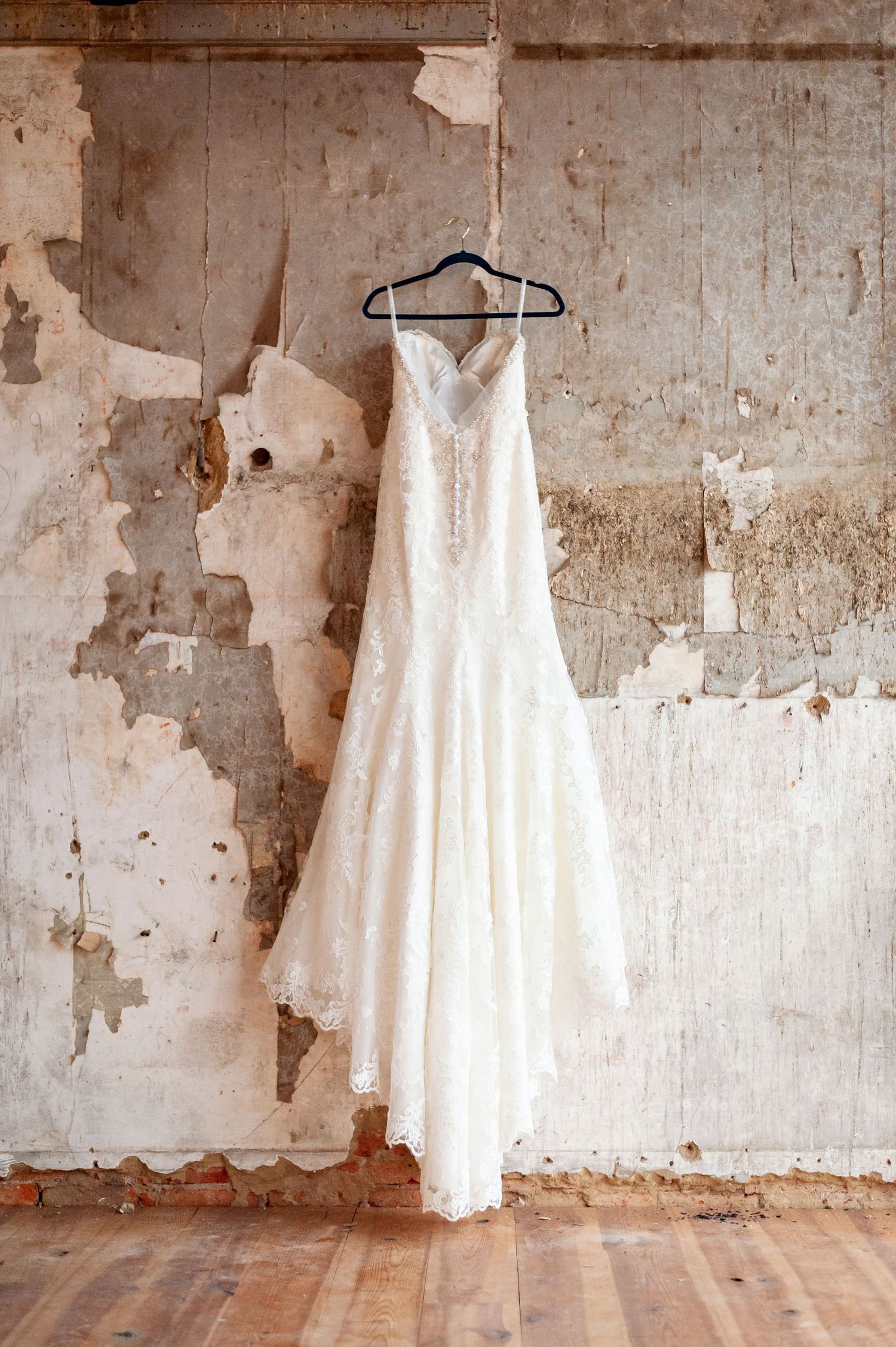 Full-length white lace wedding gown displayed against weathered industrial wall.
