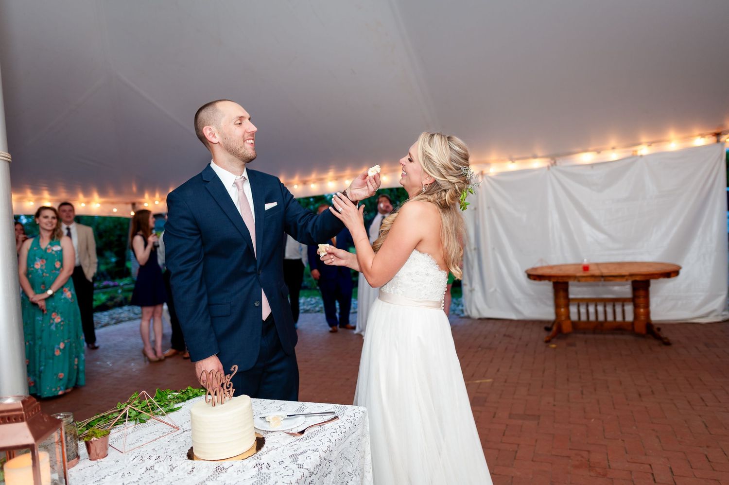 A couple sharing a playful moment while cutting their wedding cake under a tent.