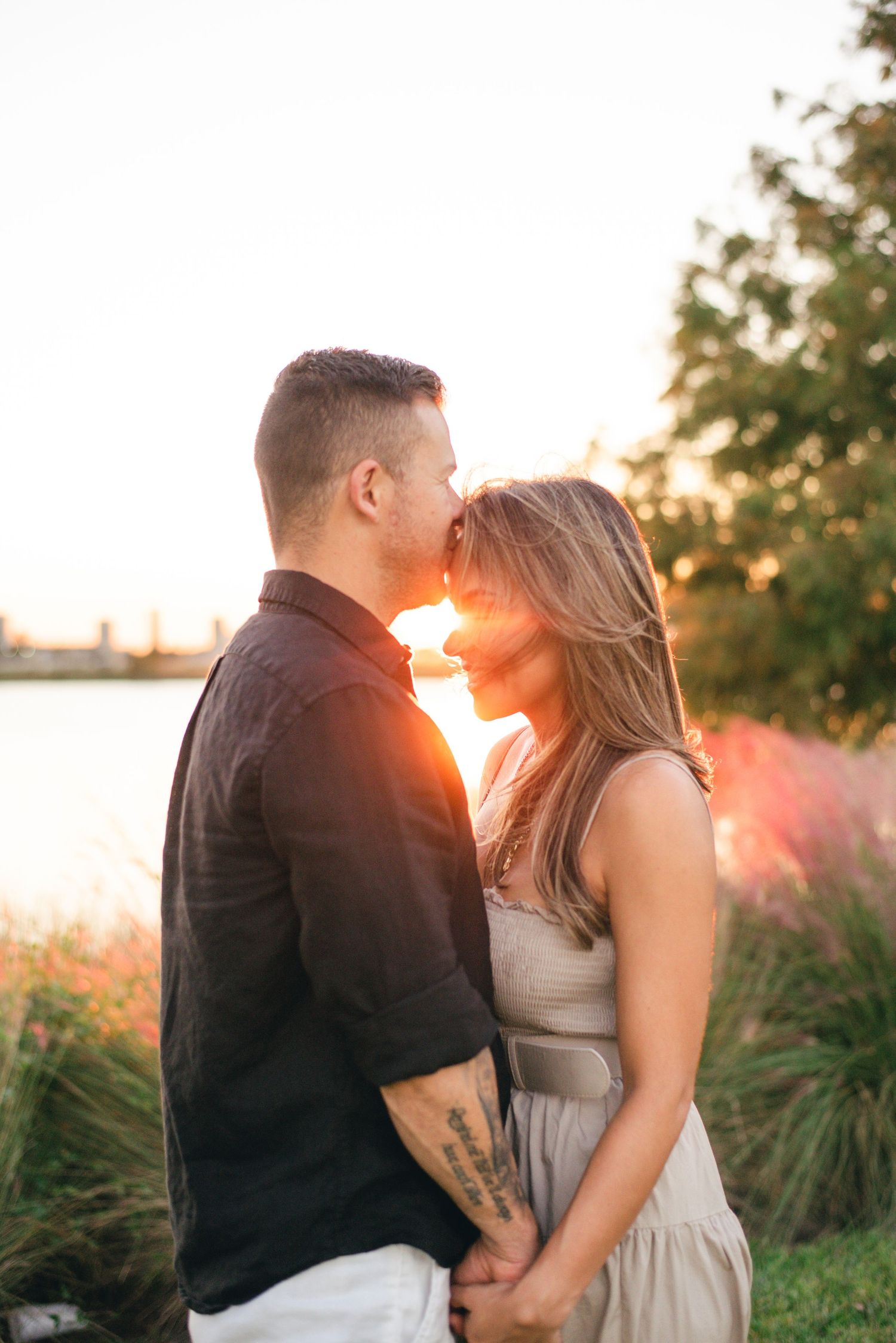 Couple embraces in tender moment with sun setting over water and ornamental grasses nearby.