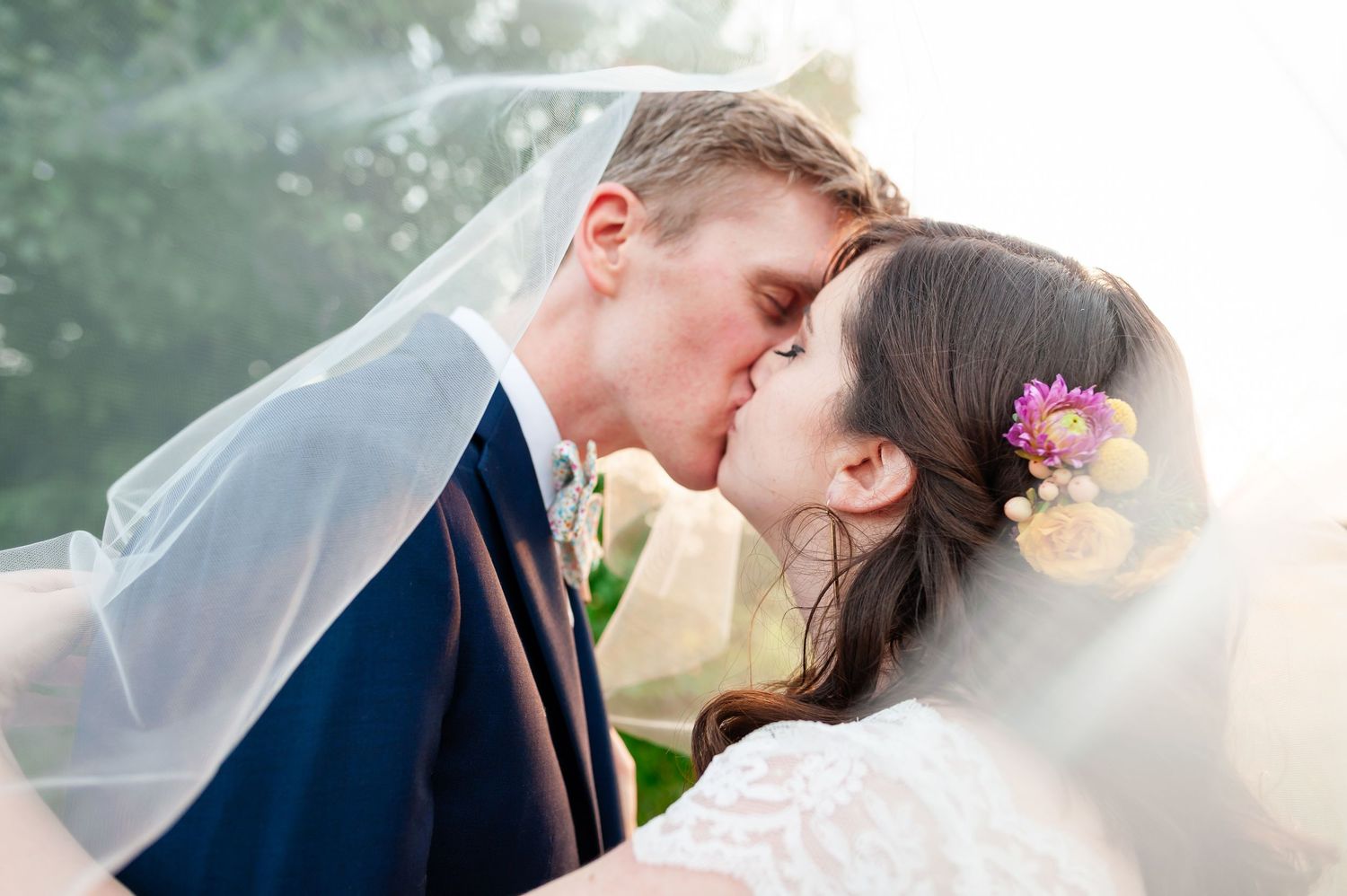 Bride and groom share intimate kiss under veil with colorful flowers in bride's hair.