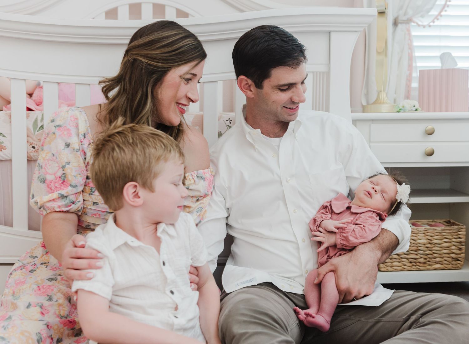 Parents and toddler sitting together smiling at newborn baby in pink outfit.