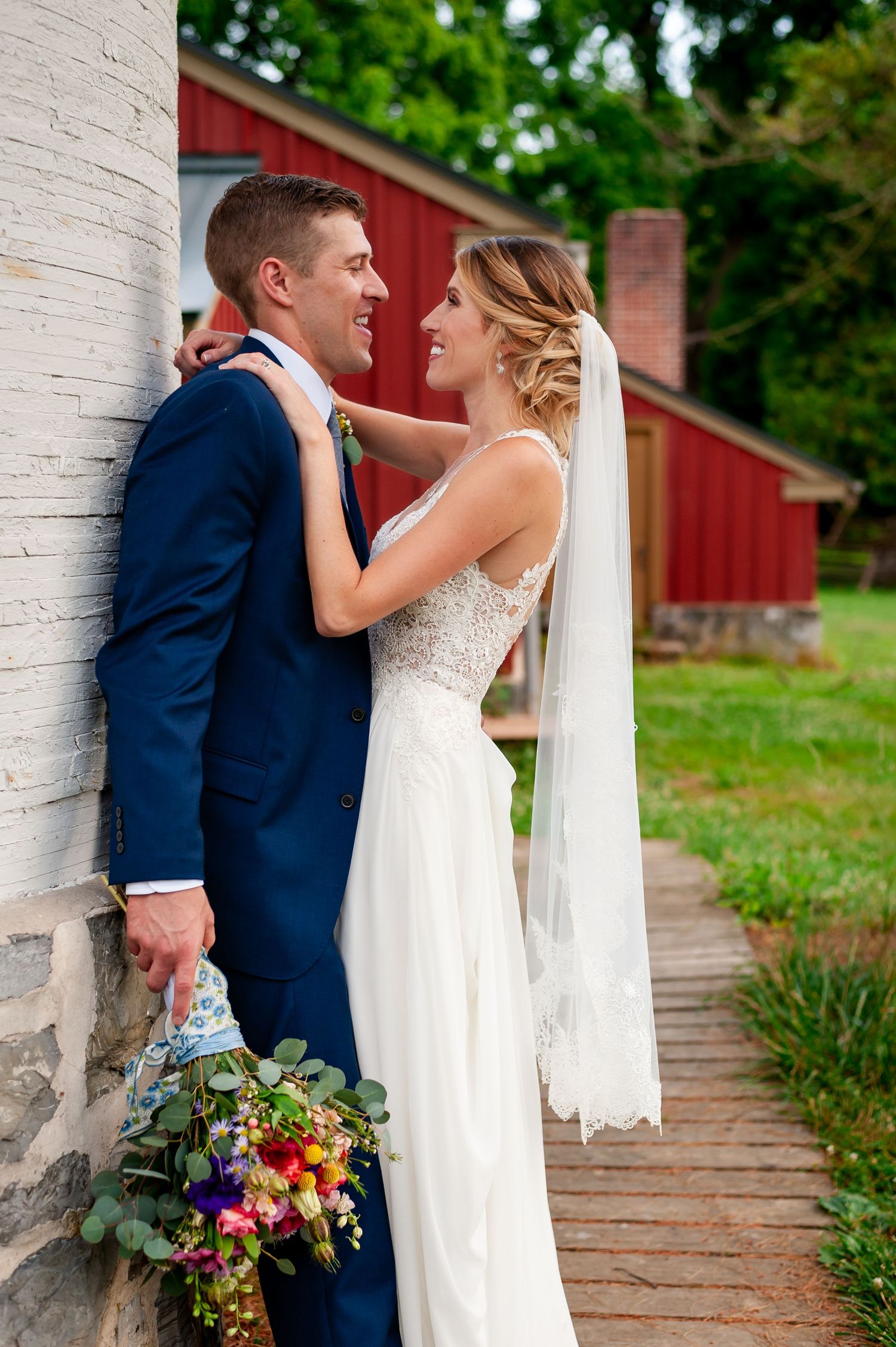 Newlyweds embrace next to a rustic red barn with bride holding a colorful bouquet.