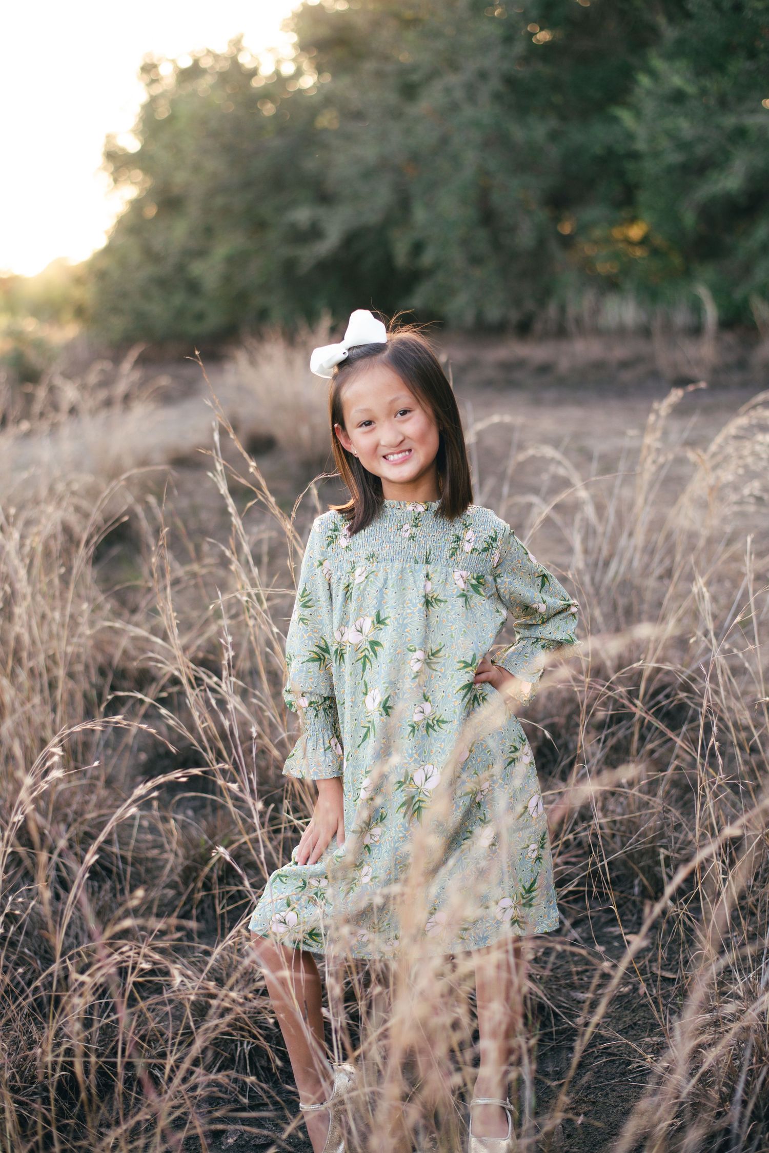 Young girl in mint floral dress stands in tall brown grass during golden hour.
