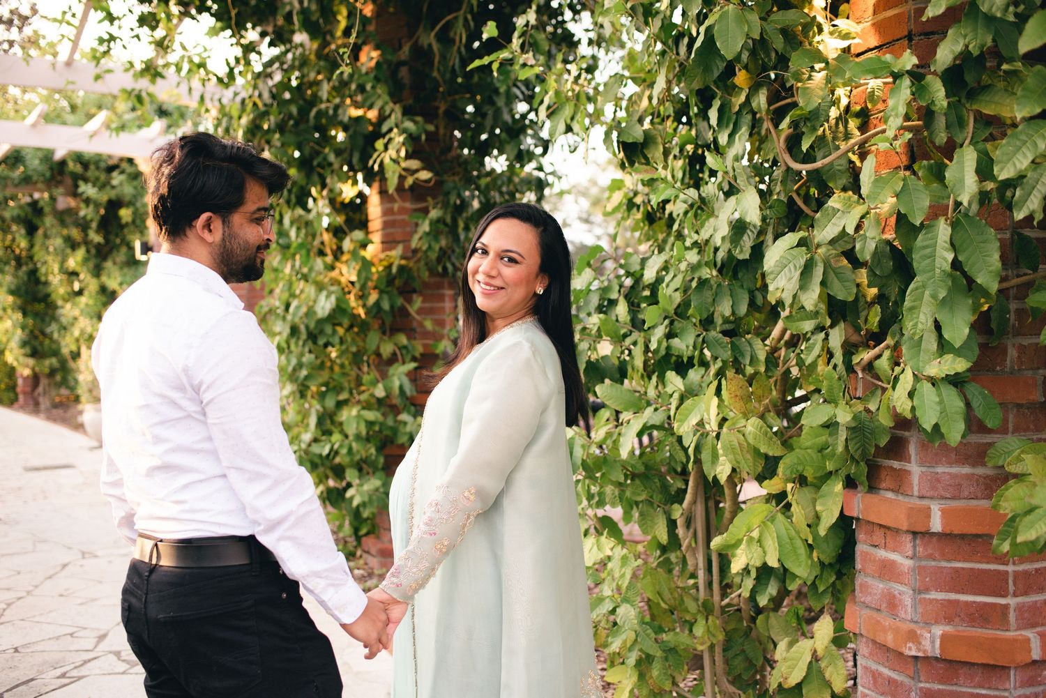 Two people hold hands and stand near a green vine-covered brick wall on a bright sunny day.
