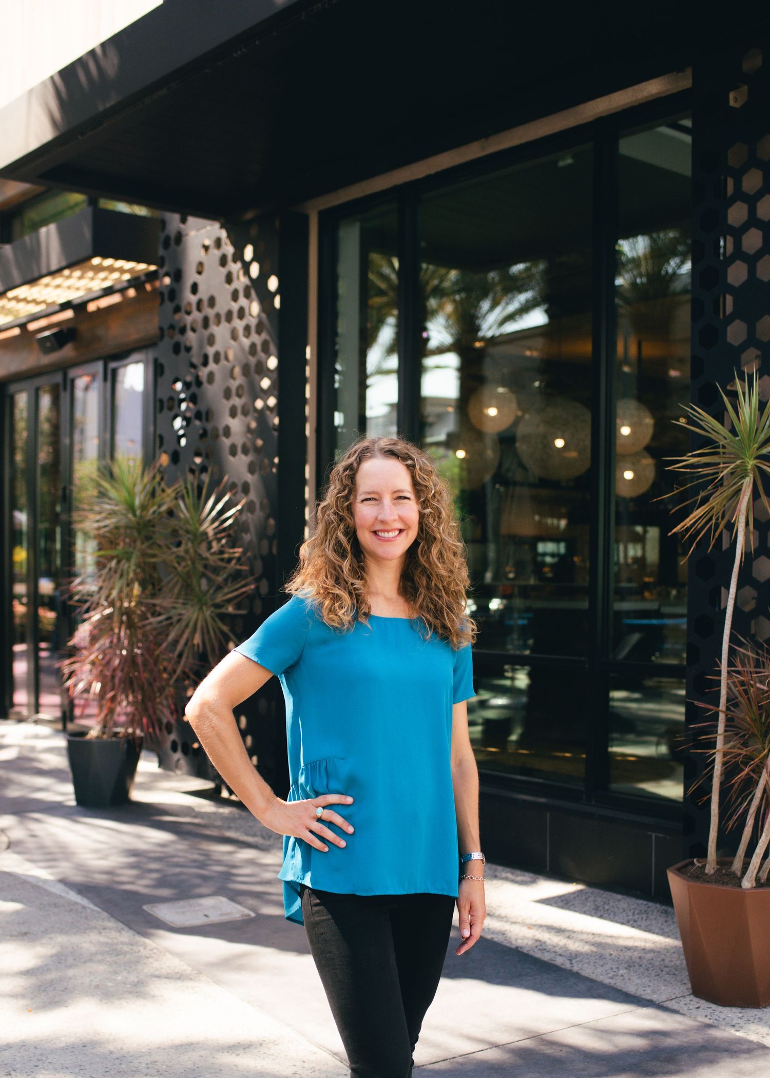 Woman in bright blue top posing against storefront with decorative lighting and plants.