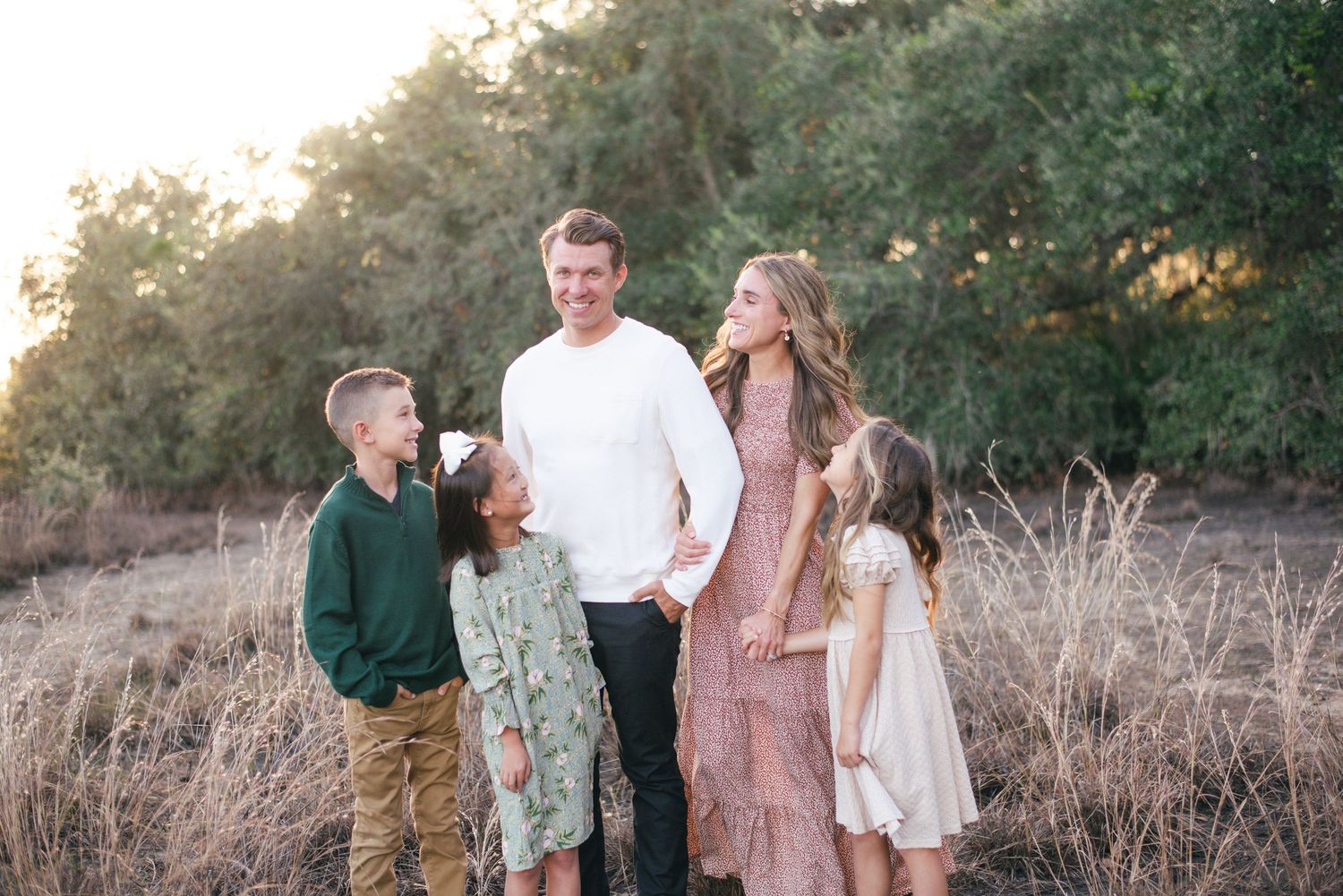 A family in coordinated neutral outfits poses for a photo session in a golden sun-lit outdoor setting.