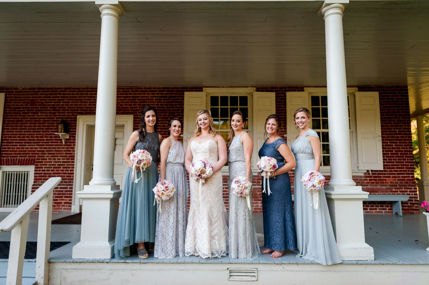 Bridal party stands in elegant formation on colonial-style porch with white pillars.