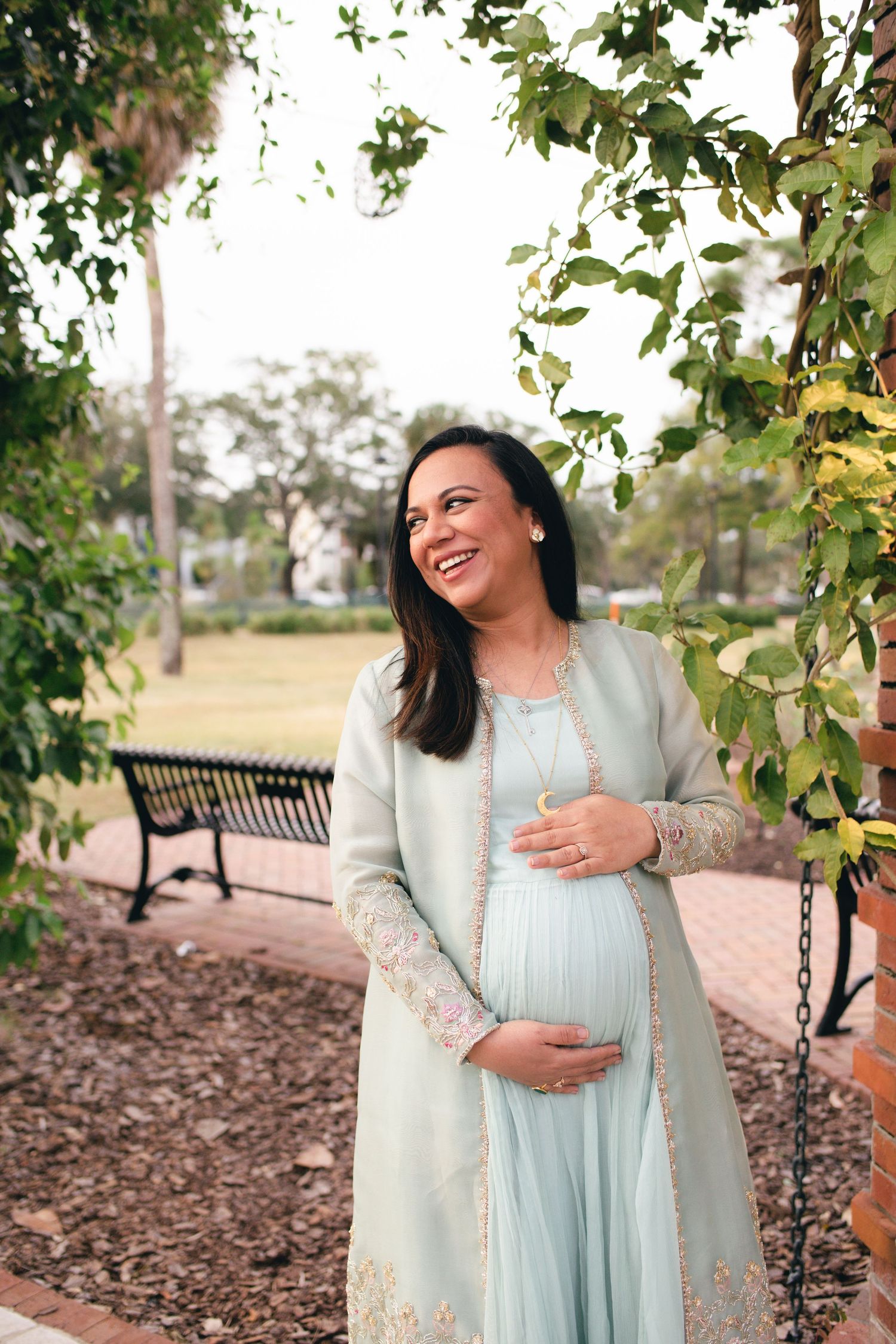 Expecting mother in a light blue dress and embroidered coat stands joyfully in a garden setting with a park bench behind her.