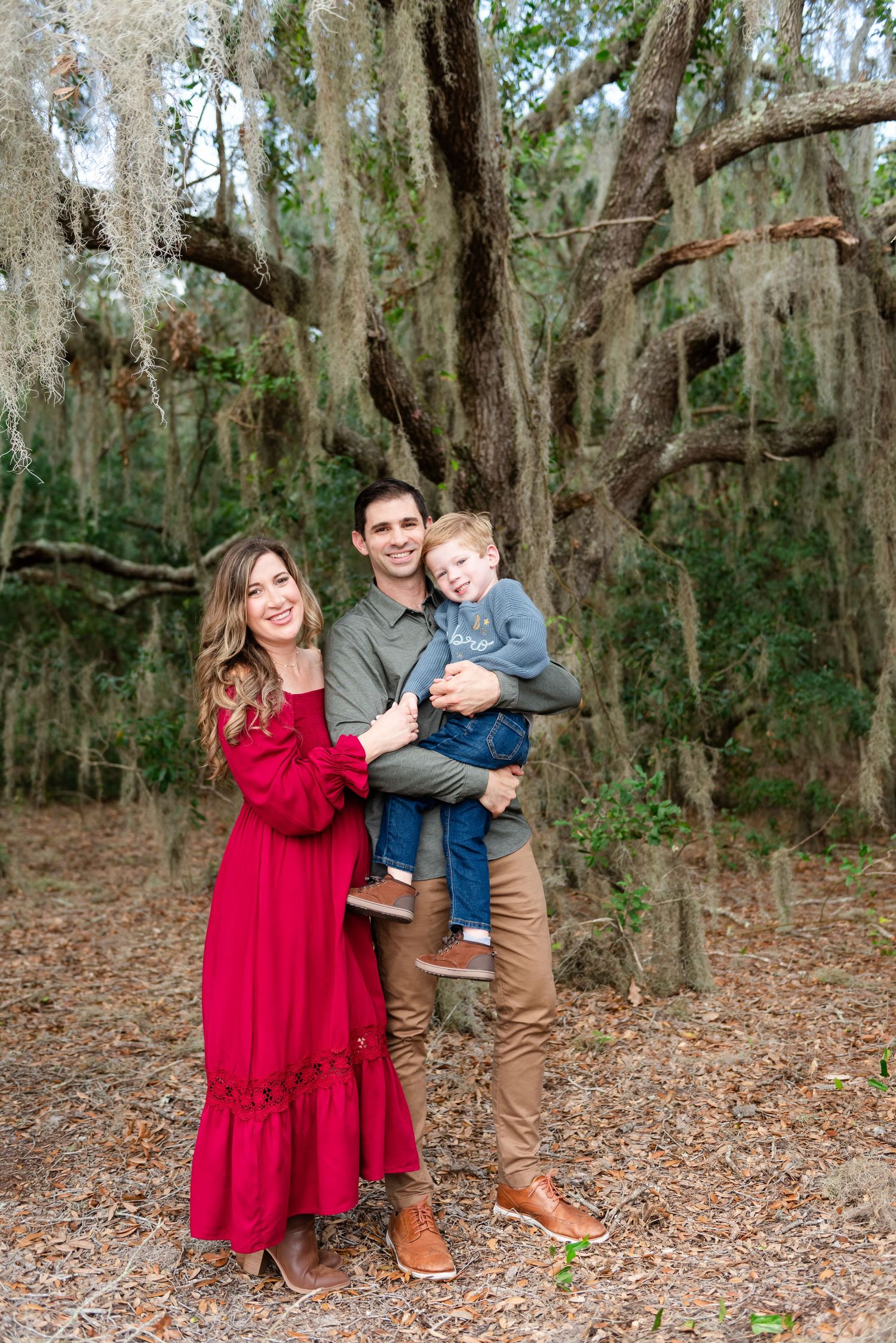 Family standing together under moss-draped oak trees, with woman in red dress and man holding small child.