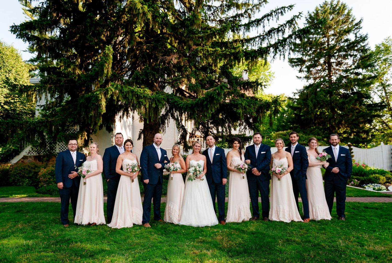 Full wedding party in navy suits and blush dresses poses outdoors on a sunny garden lawn.