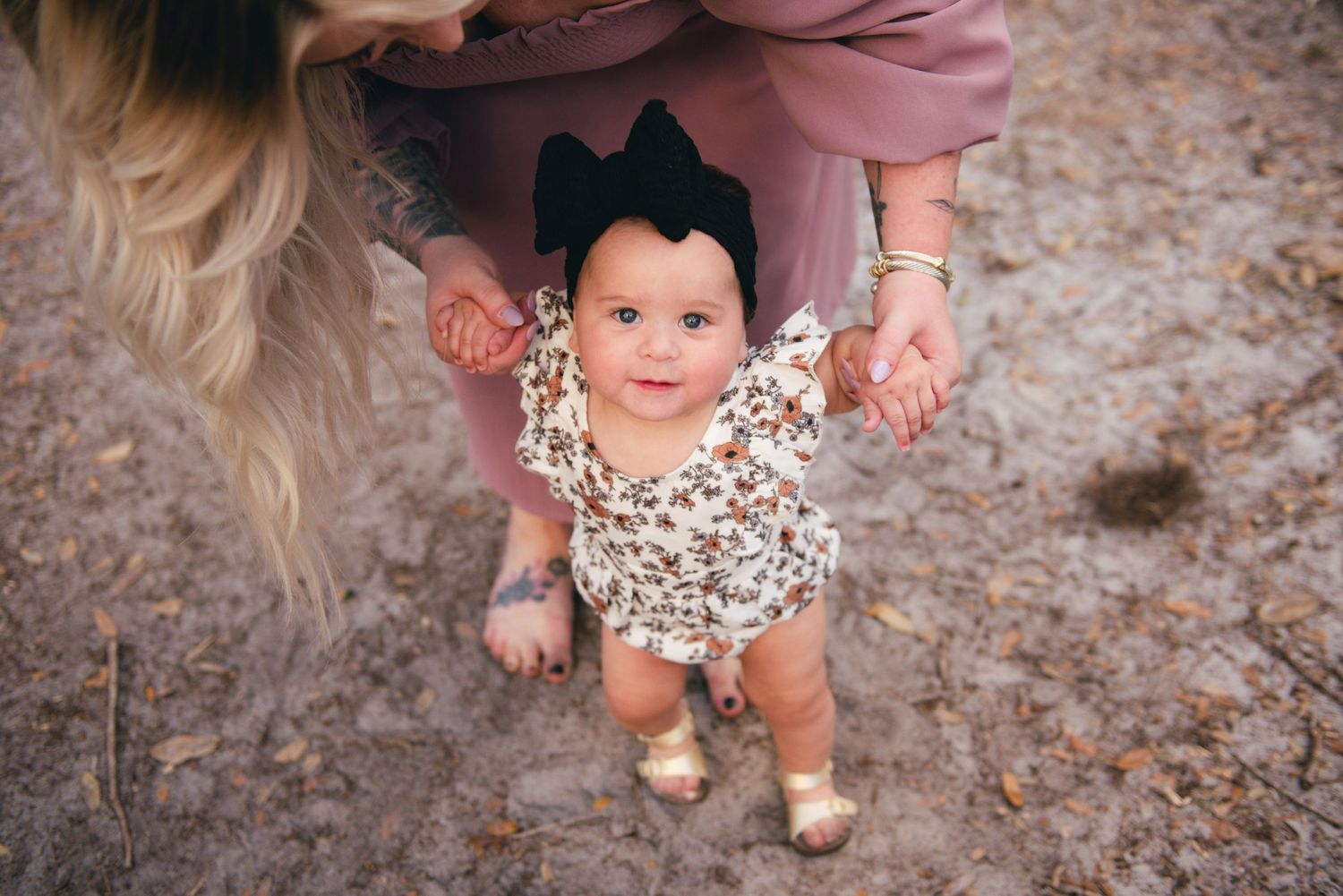 Baby in floral romper and black bow standing supported in outdoor setting.