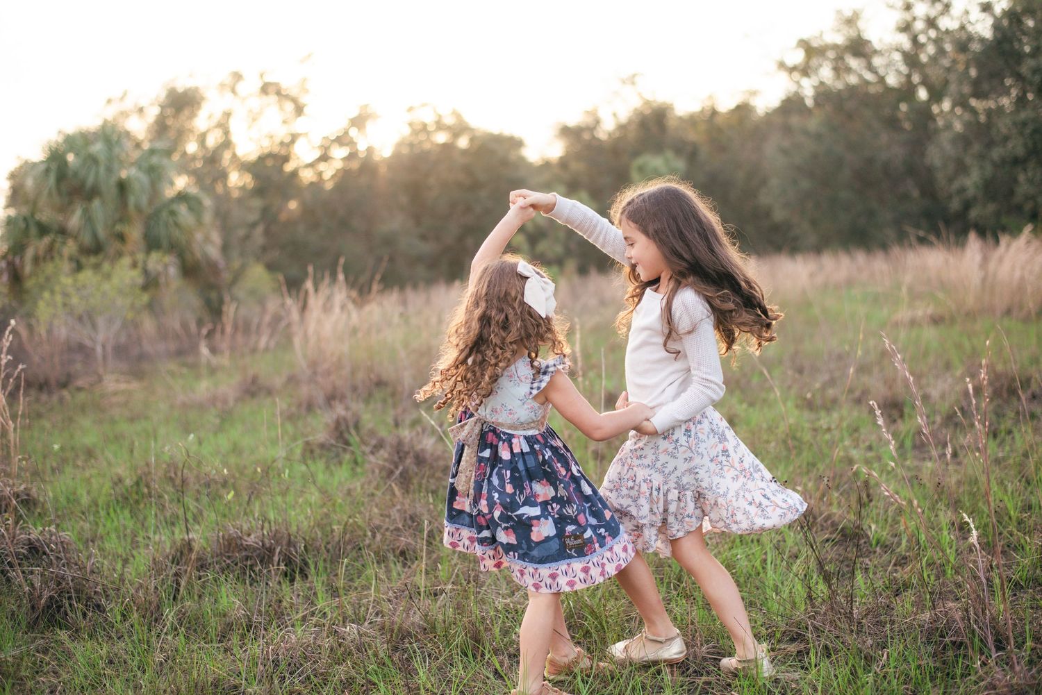 Two young girls in floral dresses dance together in a sunlit meadow at golden hour among tall grass and trees.