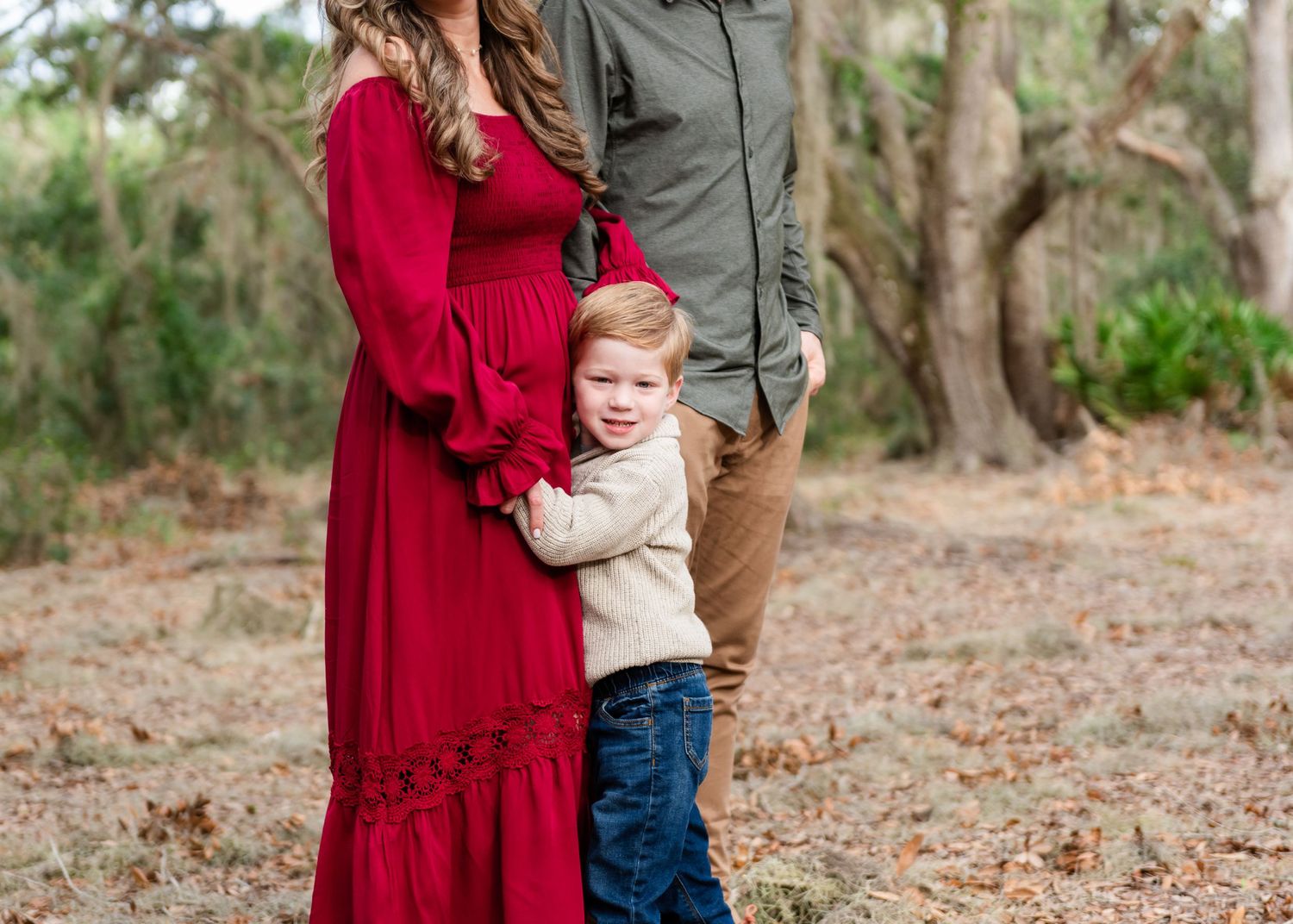 A child in a beige sweater hugs someone wearing a flowing red dress while standing among trees in a wooded area.