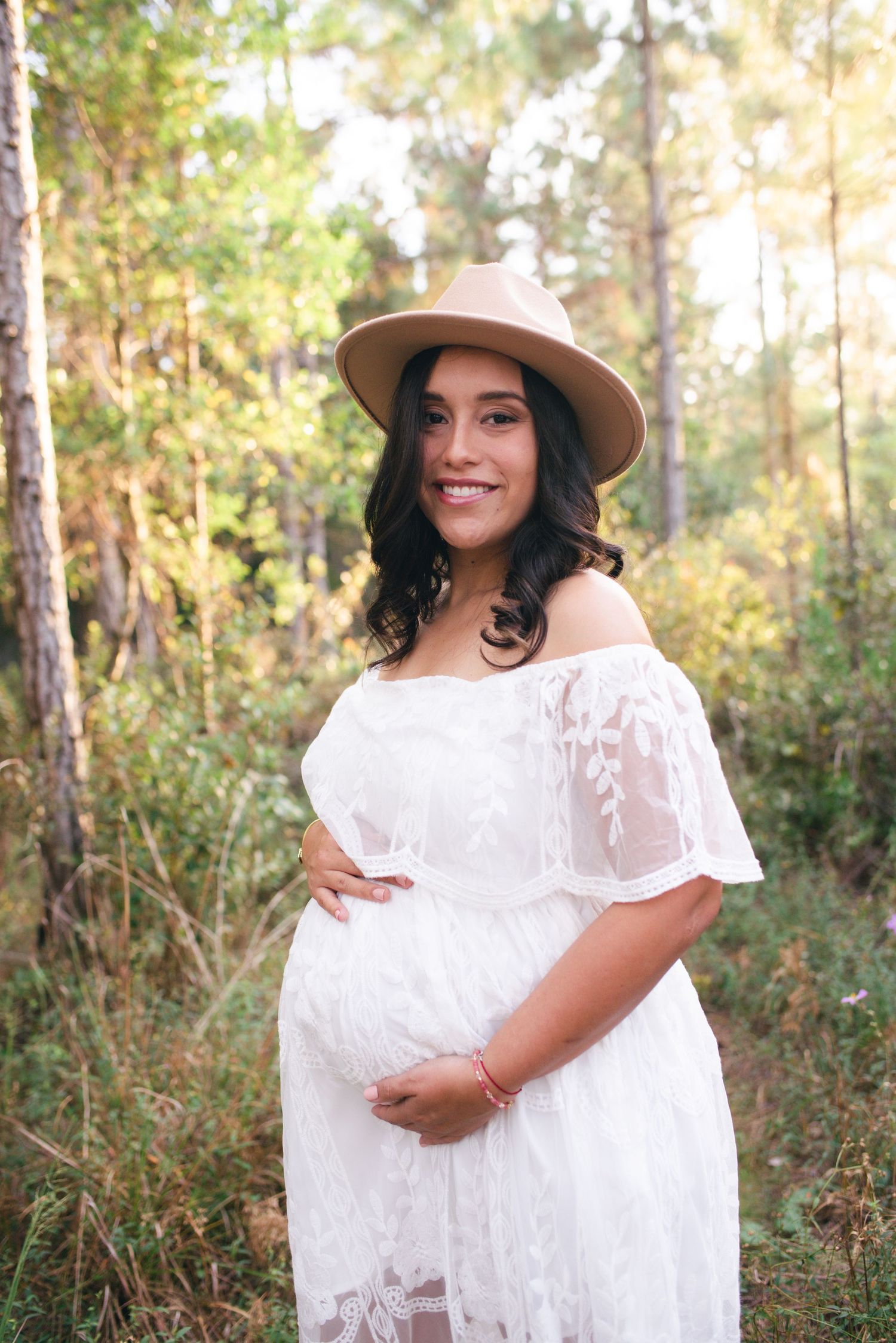 A maternity photo session in a sun-drenched forest setting shows a woman in a flowing white lace dress and beige hat.