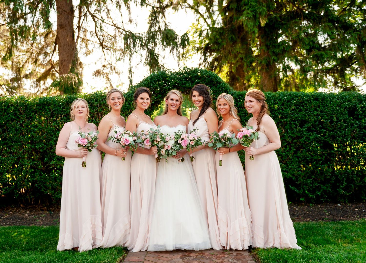 Bridal party in matching blush gowns poses on manicured lawn with hedges and trees.