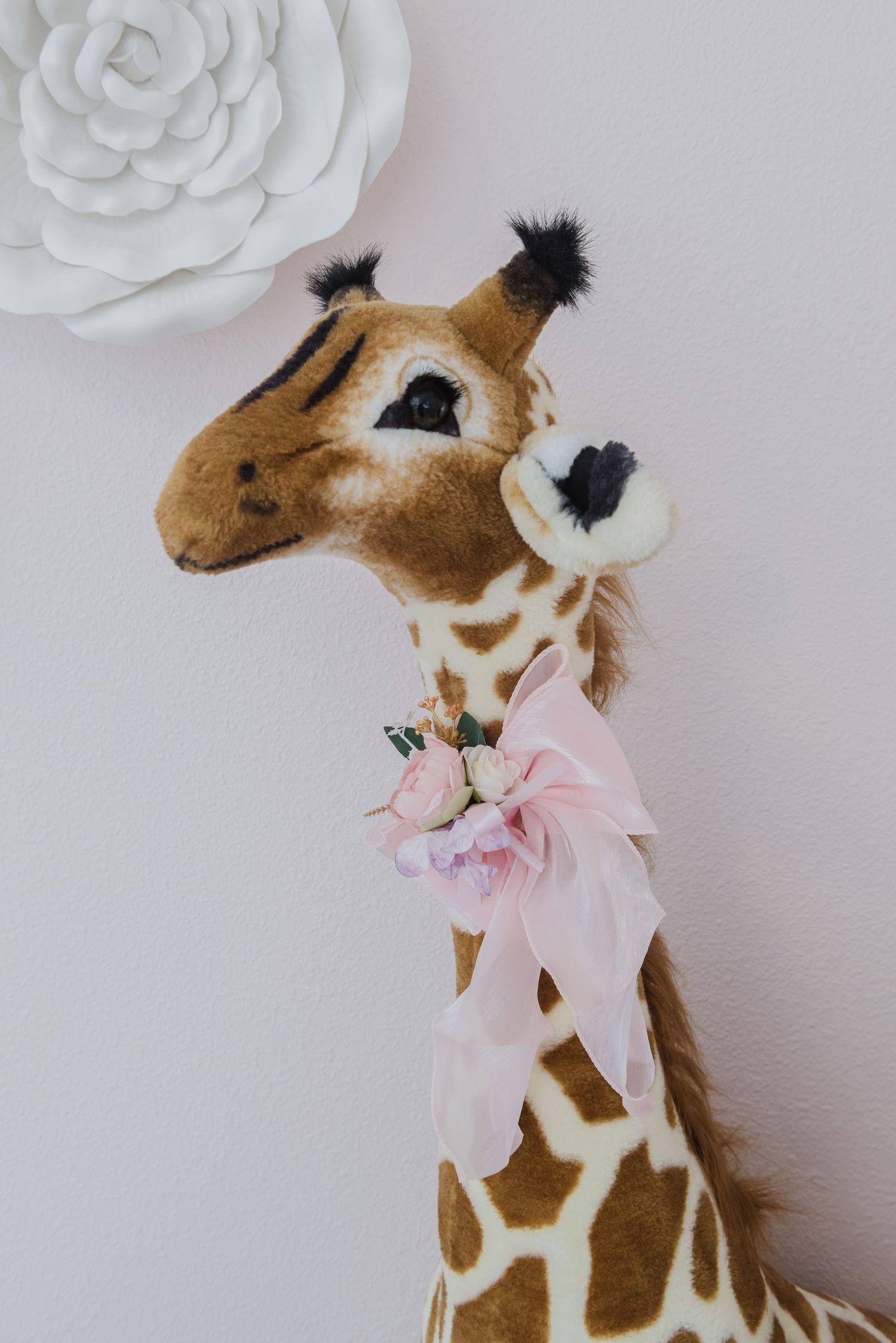 Plush giraffe with pink bow decorates nursery wall beneath white paper flower.