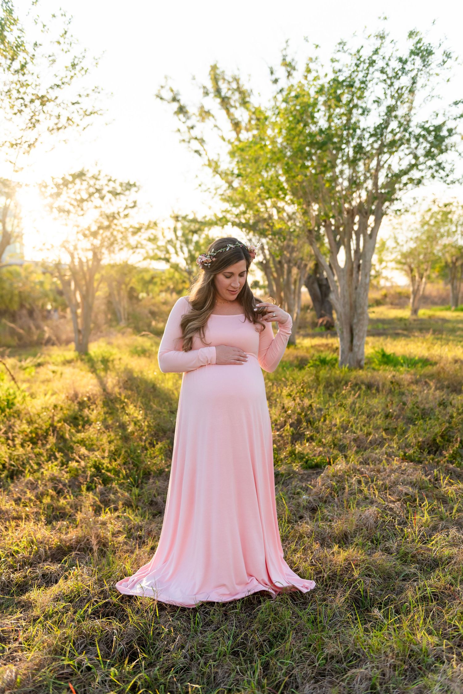 A pregnant woman in a flowing pink maxi dress stands in a grassy field at golden hour with trees in the background.