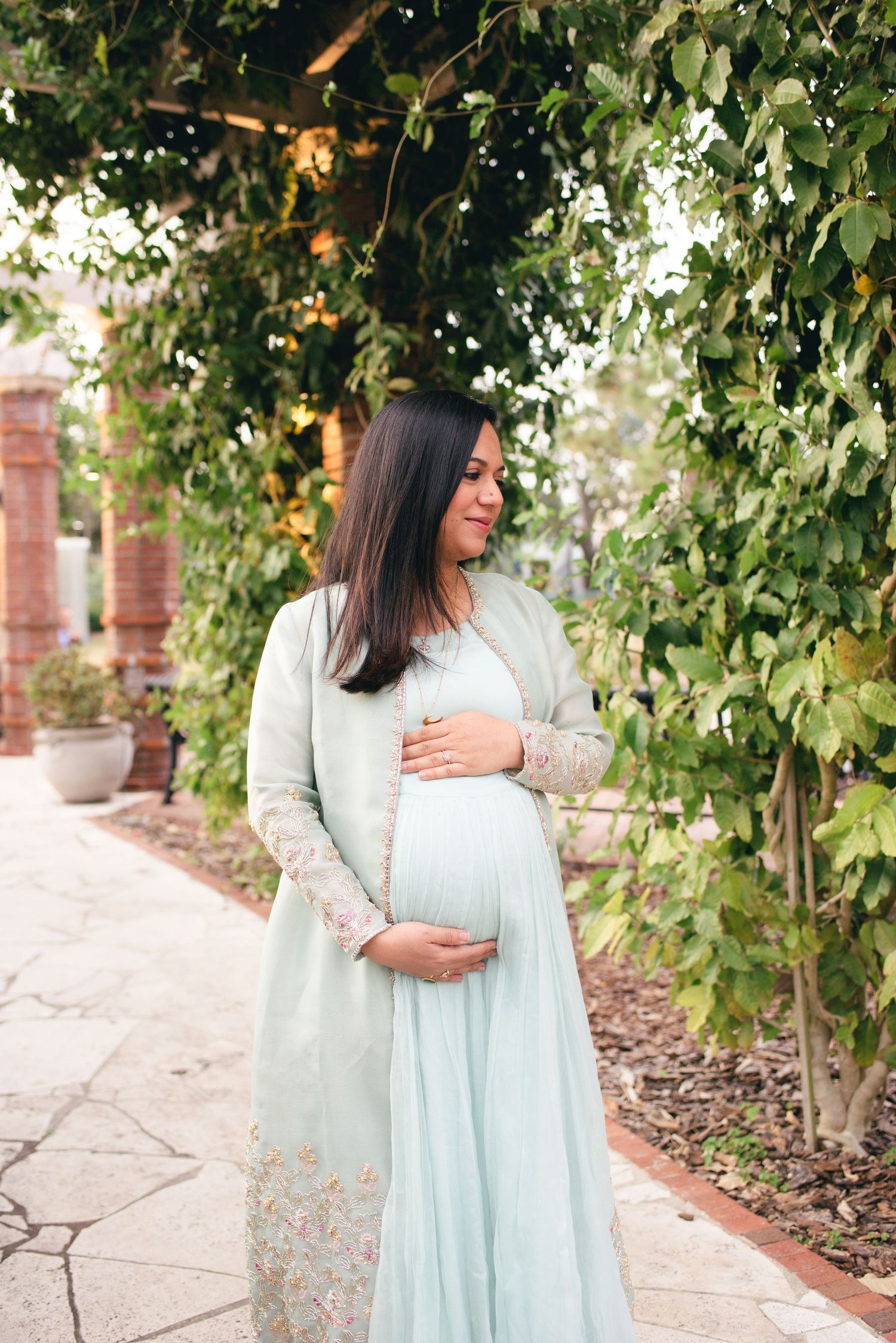 A pregnant woman in a light blue dress and cream cardigan stands beneath a leafy arbor in a garden.
